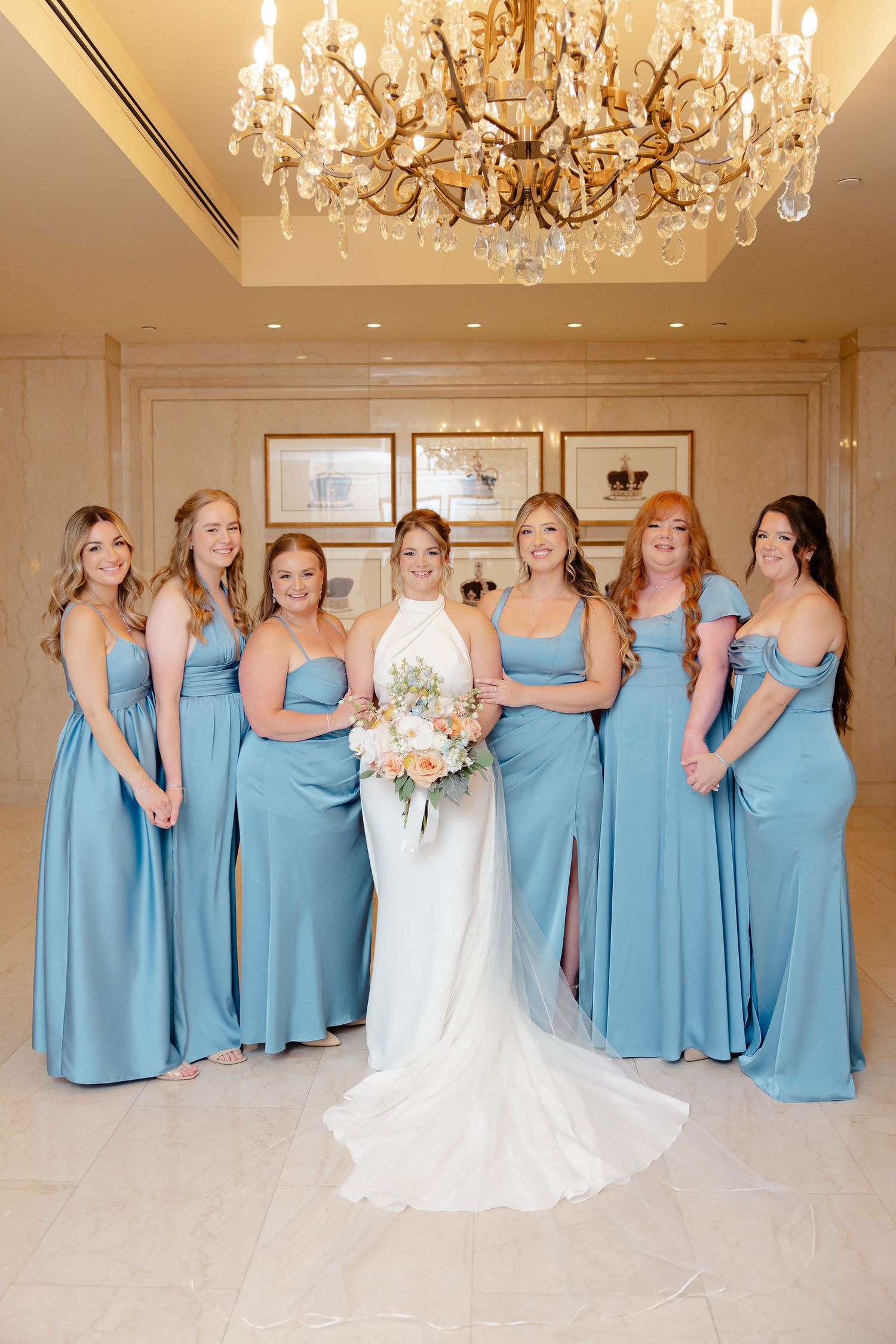 Wedding party poses in a grand ballroom with bridesmaids in light blue dresses and bride in white gown under chandeliers.