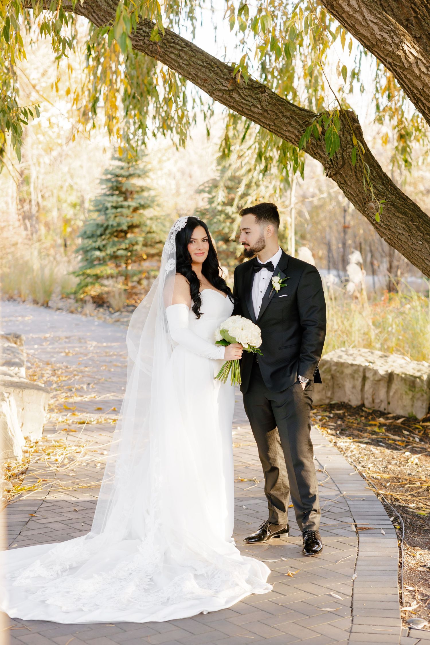 Wedding couple stands on stone pathway under dramatic willow tree branches.
