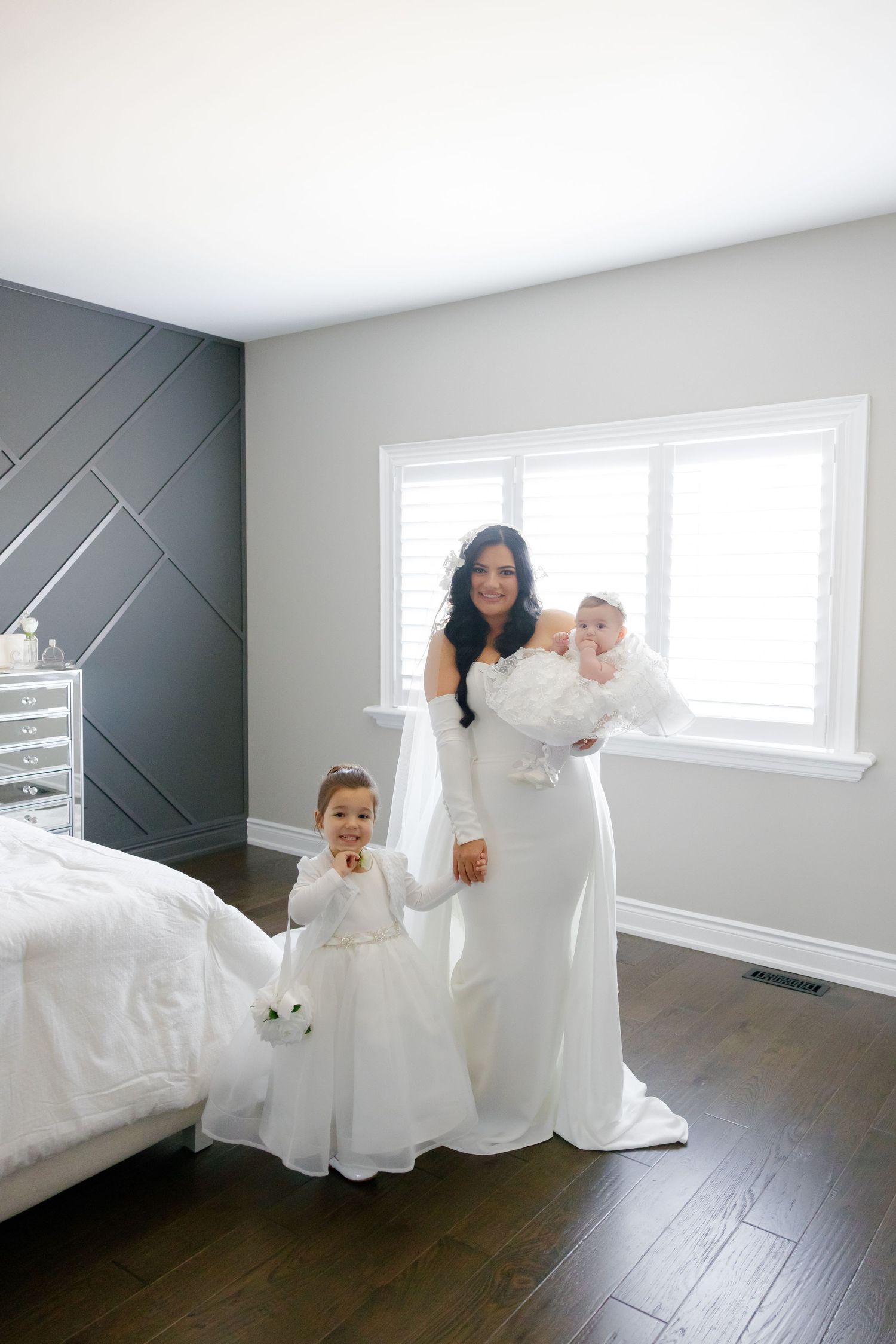 A bride in white gown poses with two flower girls wearing matching dresses in a bright room.