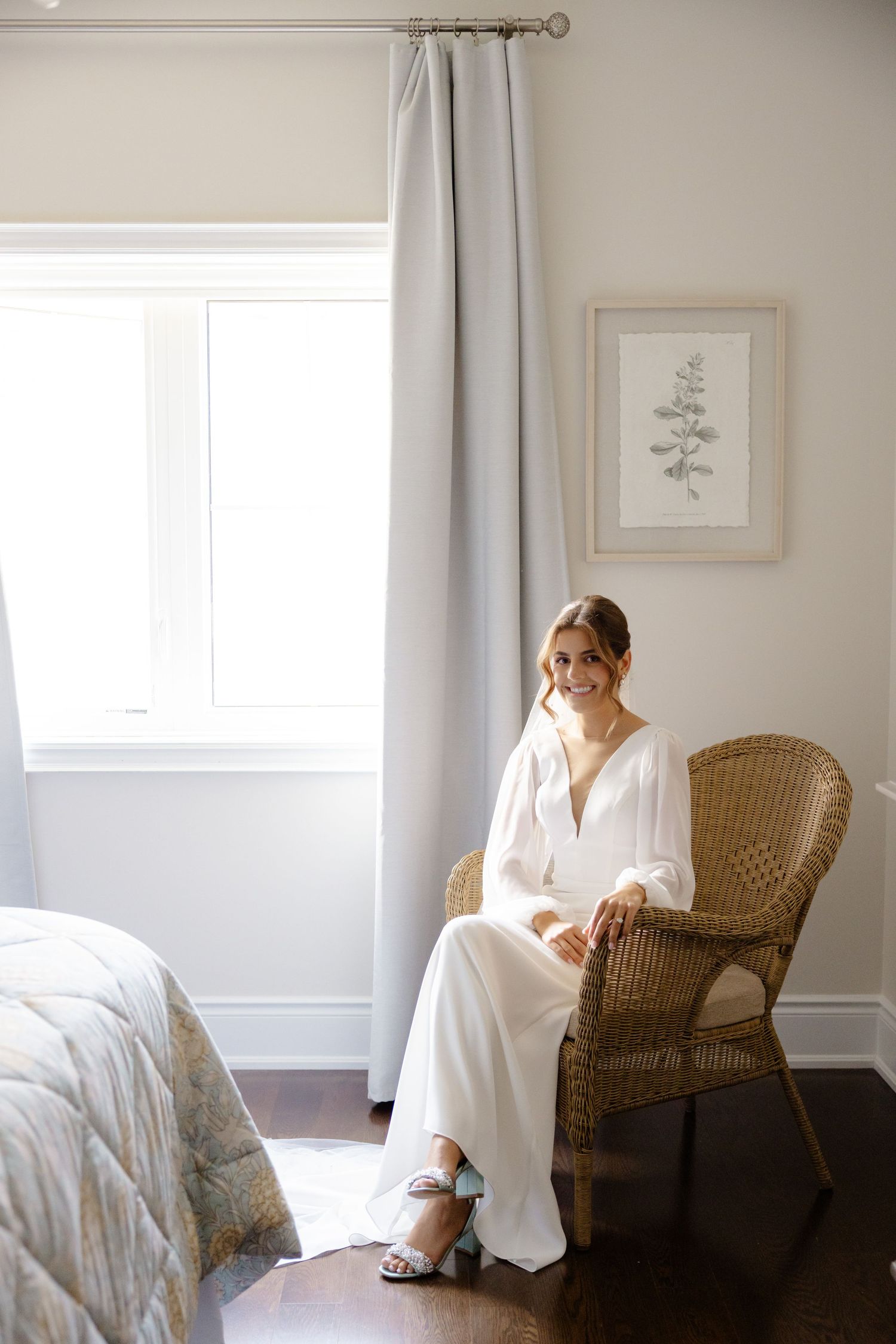 Natural light streams into bedroom as bride sits peacefully in wicker chair before ceremony.