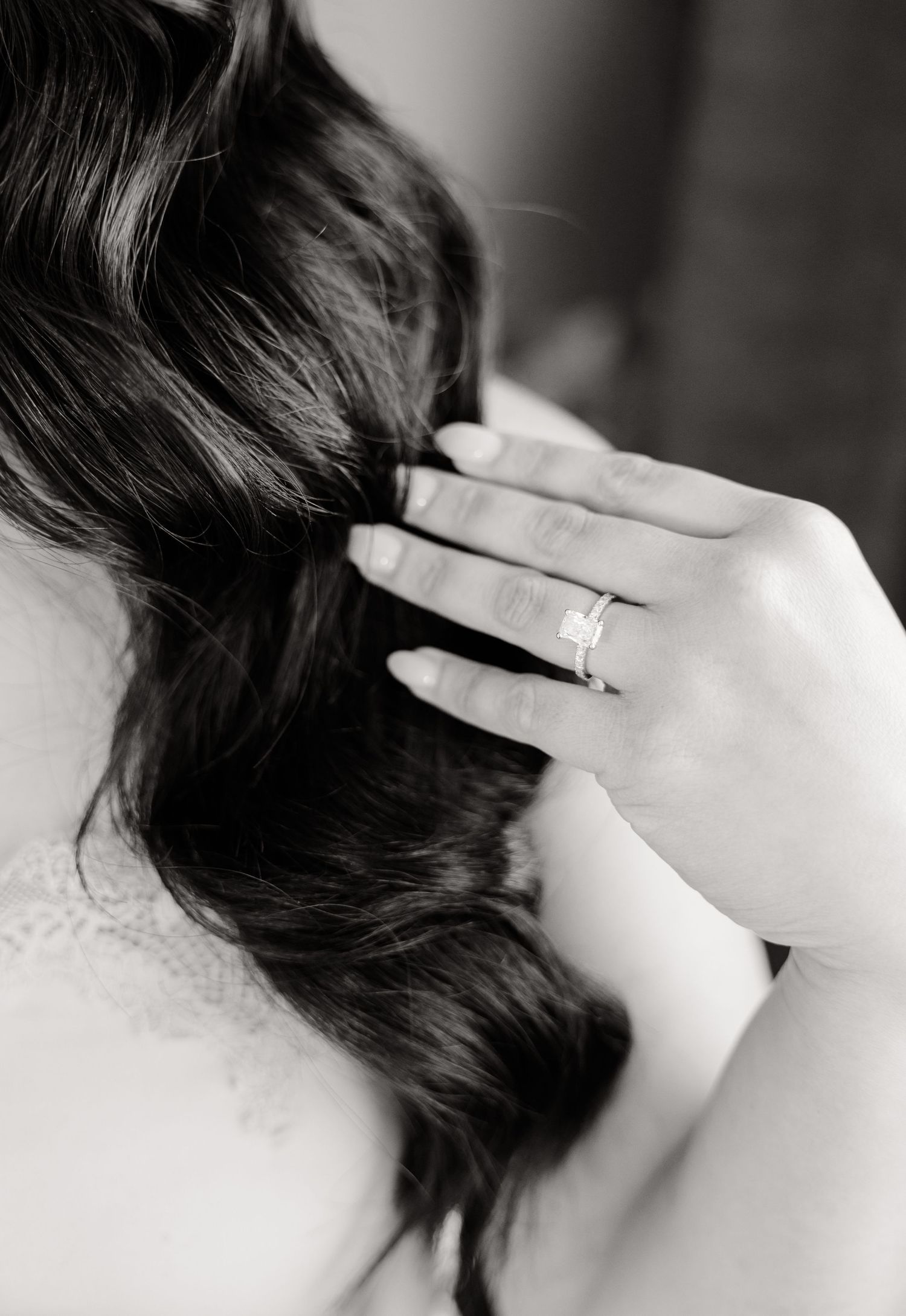 Close-up black and white photo of hands touching long dark hair, showing an engagement ring.