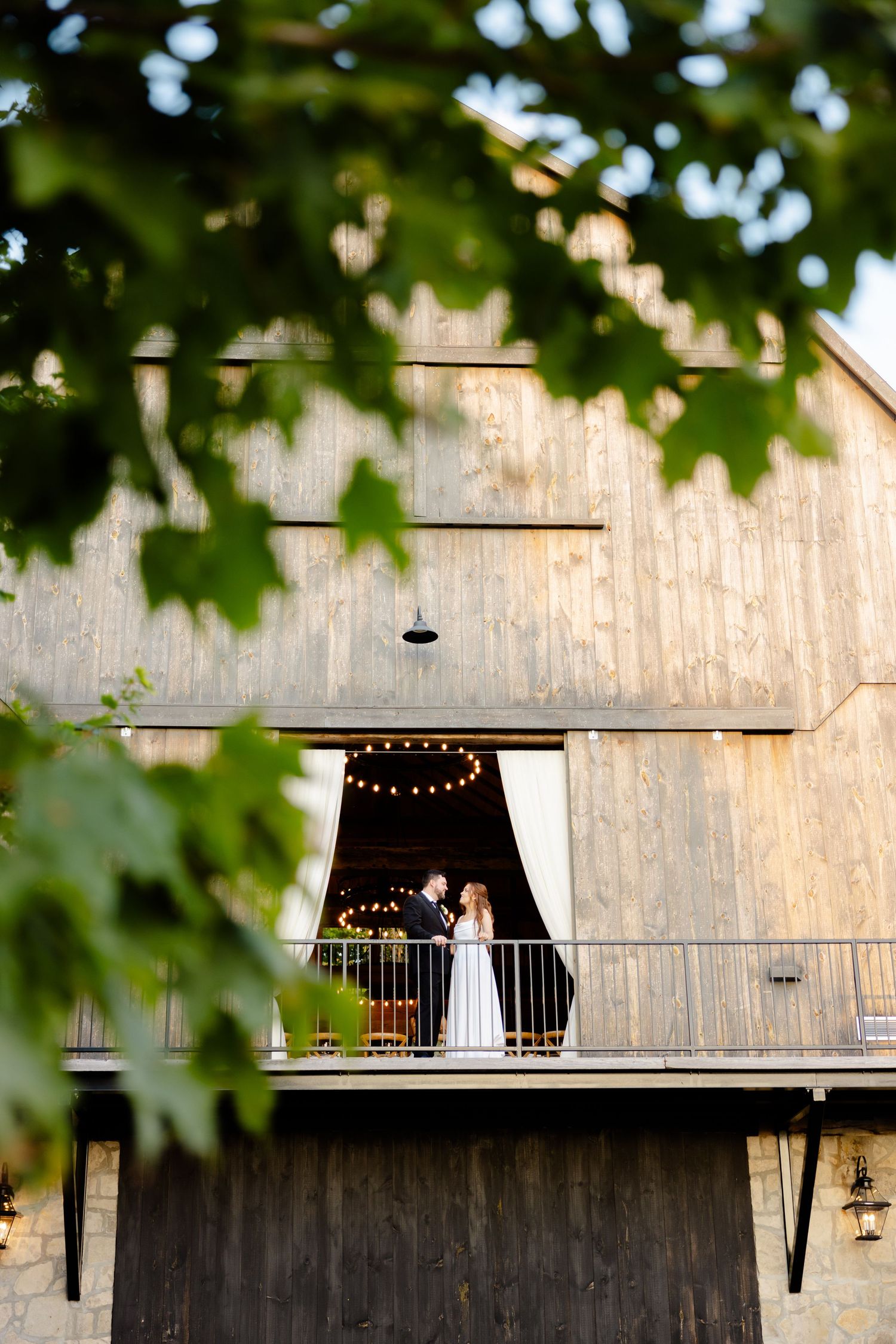Wedding scene framed by leaves showing couple on rustic barn balcony with string lights.