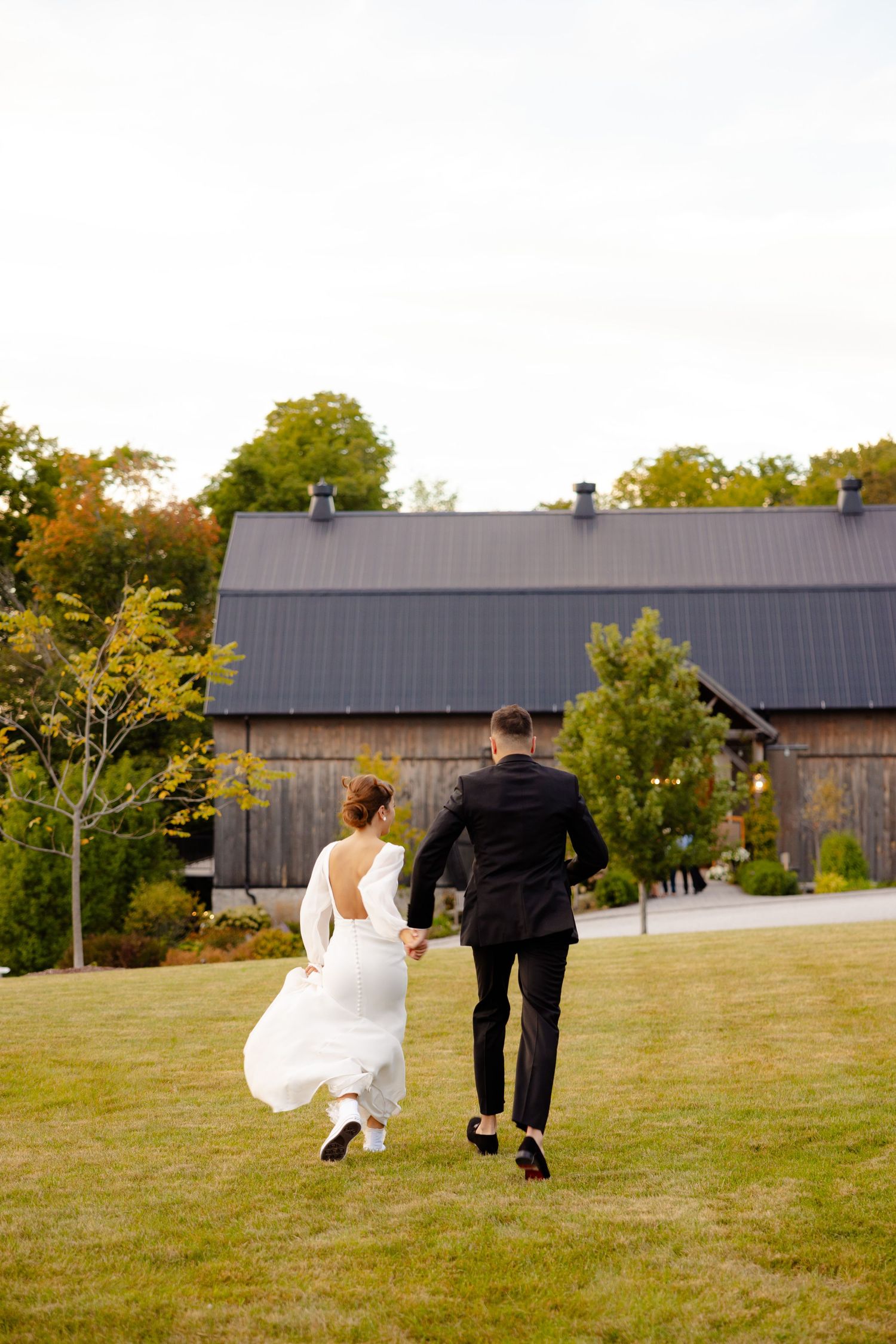 A couple runs across a lawn towards a rustic barn during their wedding celebration.