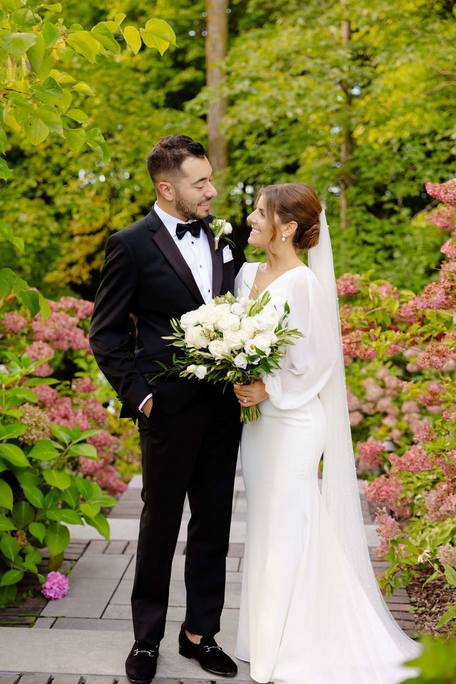 Romantic wedding photo of couple exchanging loving glances on garden path with pink florals.