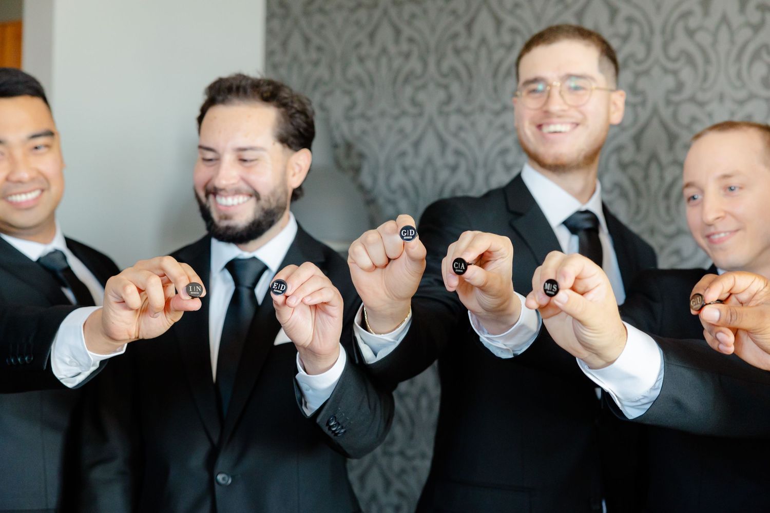 A group of groomsmen in black suits showing off silver rings on their fingers against a patterned wallpaper background.