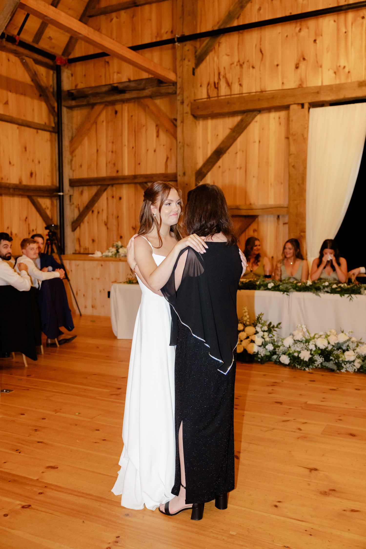 A couple shares their first dance on a wooden floor in a barn with guests watching from tables.
