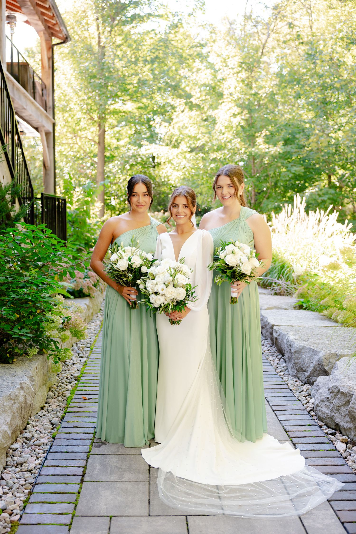 Wedding party in white and sage green dresses pose on stone garden pathway with bouquets.