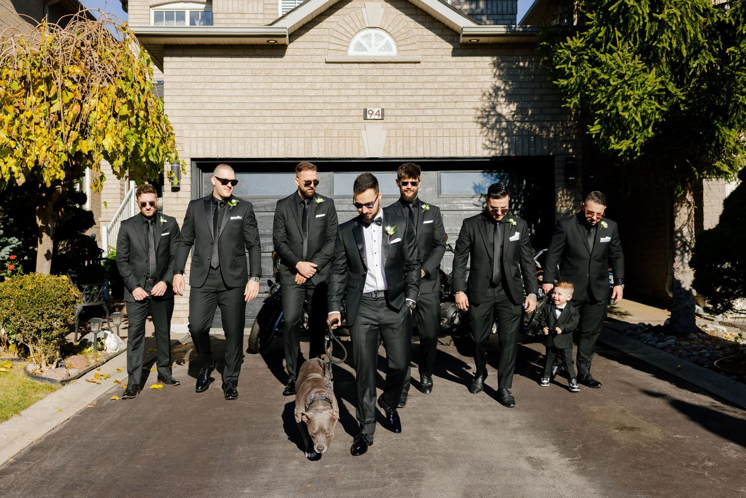 People in dark suits walking with a playful dog on a suburban driveway.