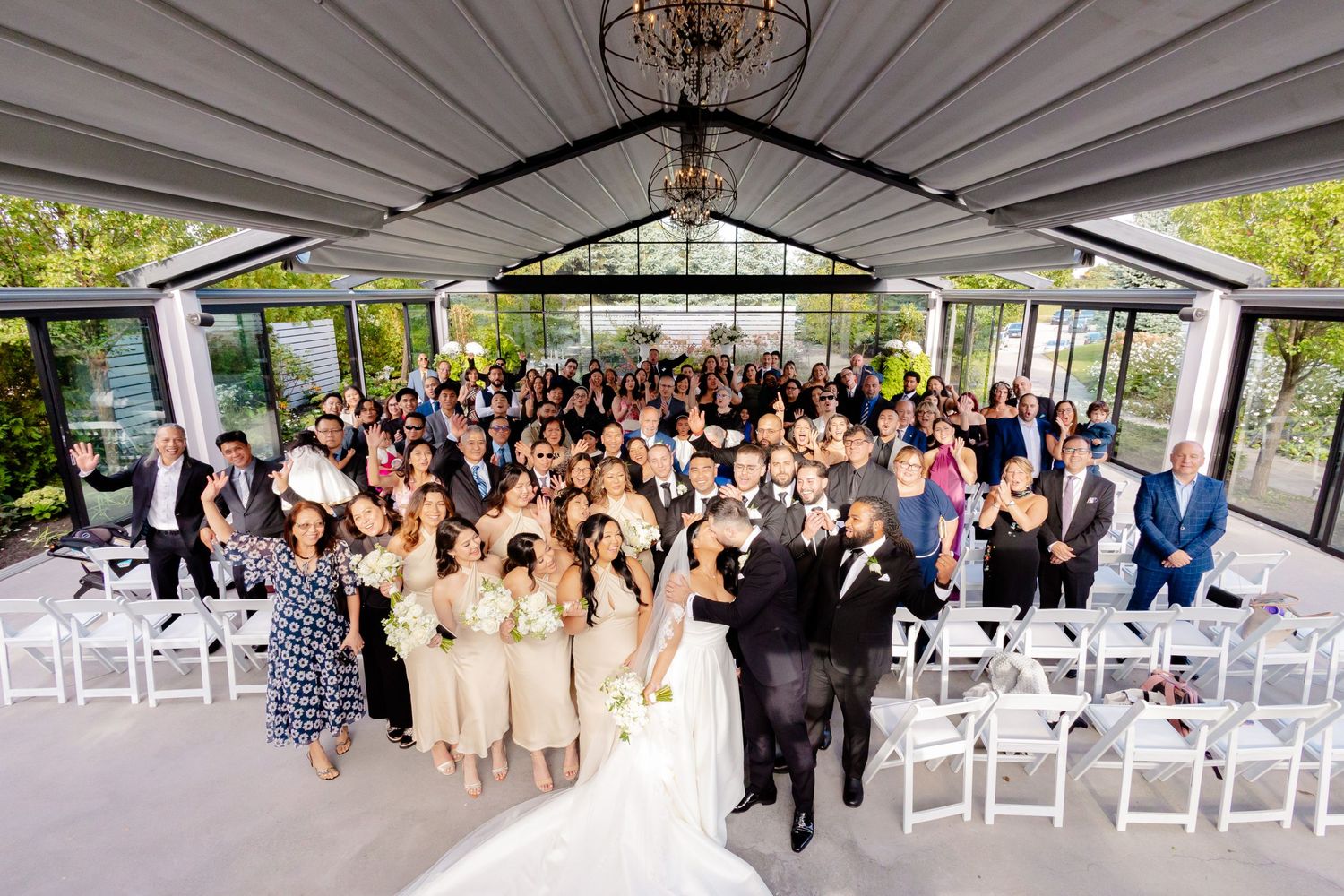 Aerial view of wedding guests gathered for a group photo in a modern glass-enclosed venue.