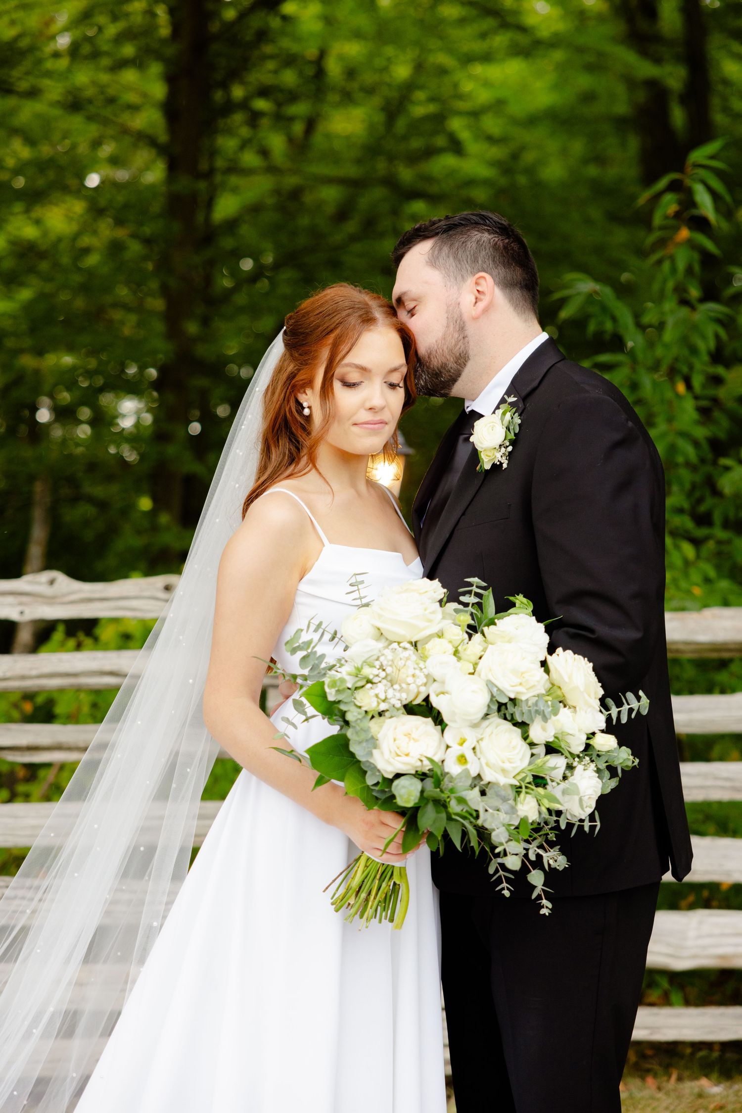 Tender moment between newlyweds with beautiful white floral bouquet in a forest setting.