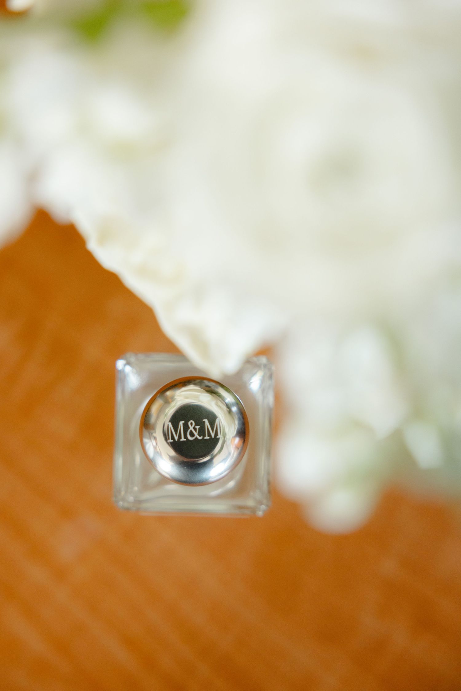 A close-up detail shot of an M&M monogram on a glass perfume bottle against a wooden surface.