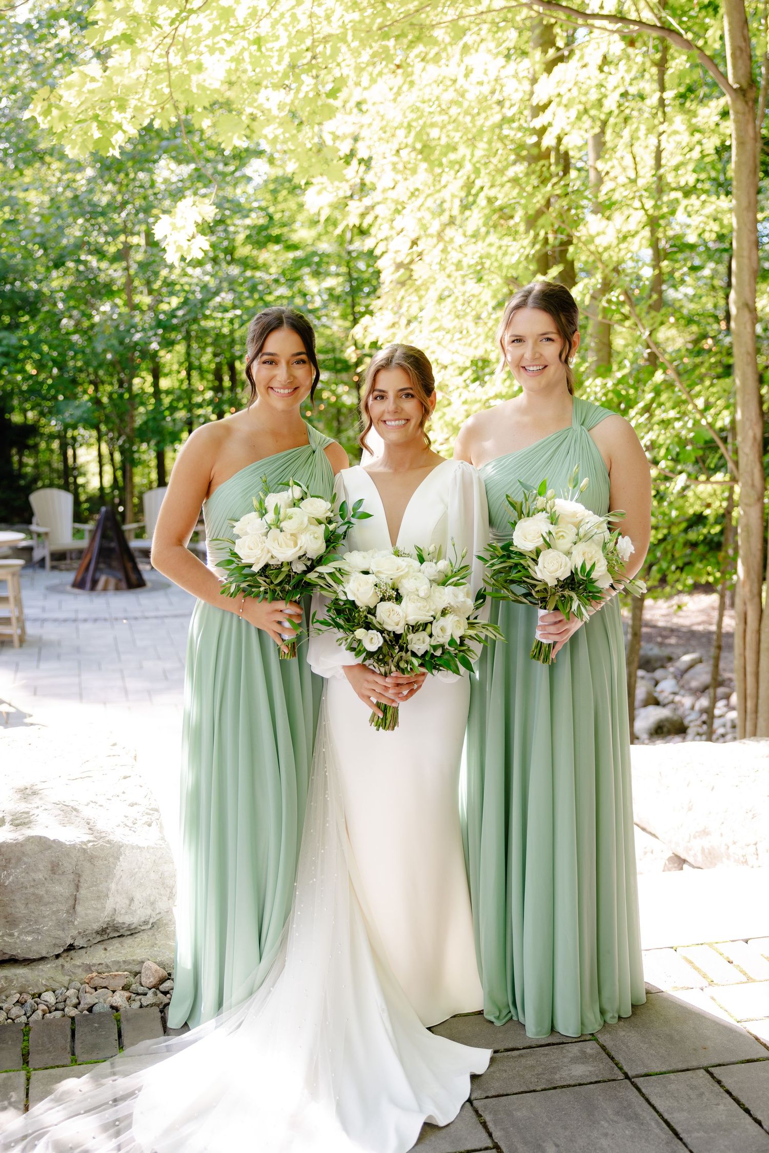 Wedding party in sage green bridesmaid dresses and white gown pose with matching white bouquets outdoors.