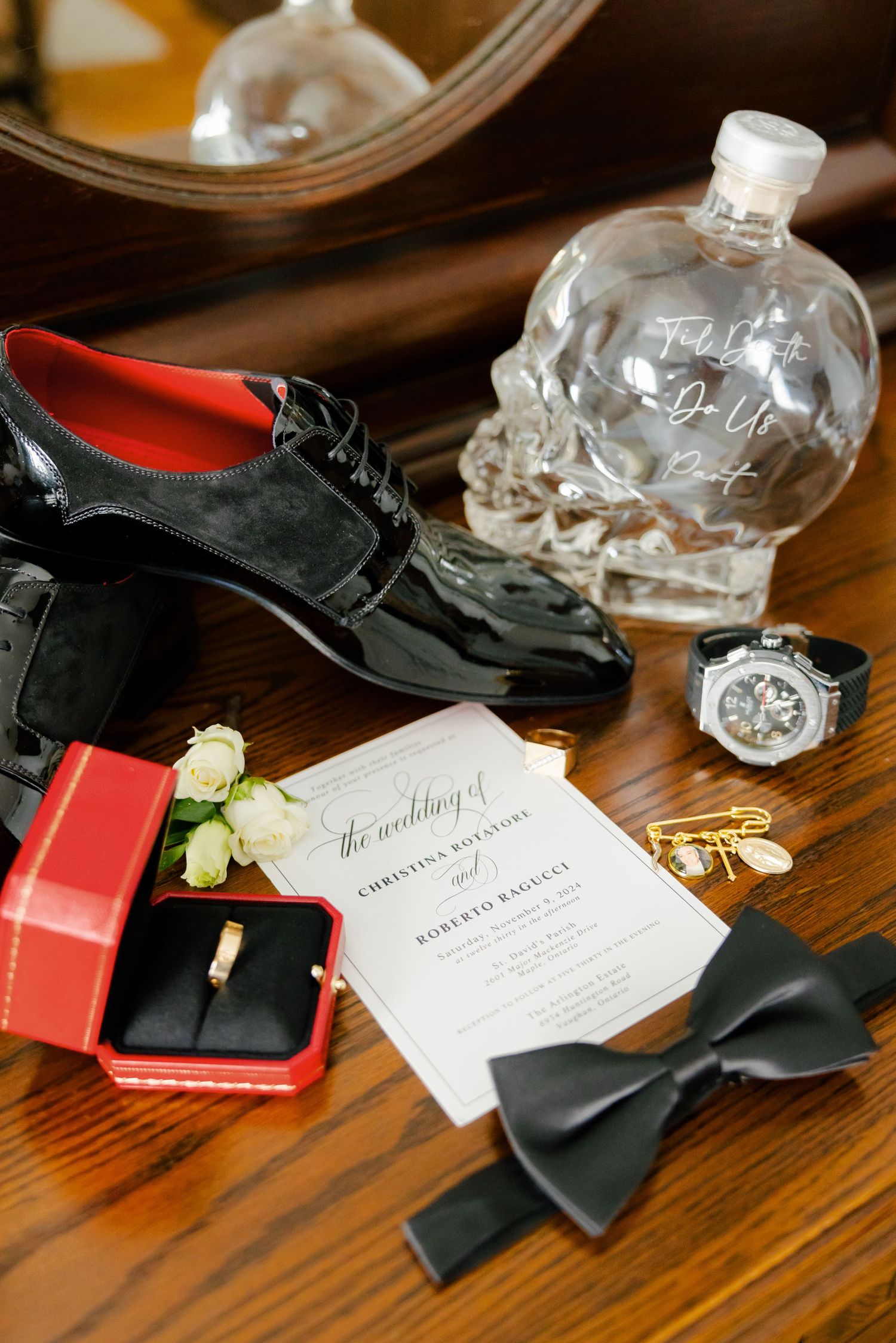 Wedding accessories including dress shoes, crystal skull bottle, bow tie and ring box on wooden table.