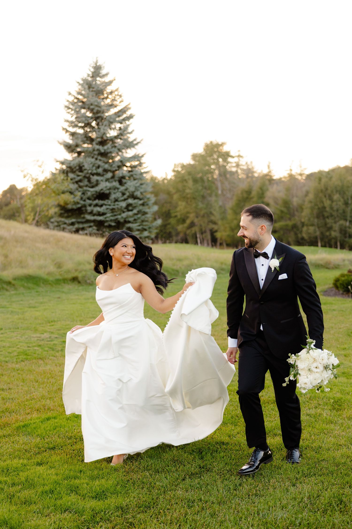 Joyful wedding couple walking on lawn with white dress billowing in the breeze.