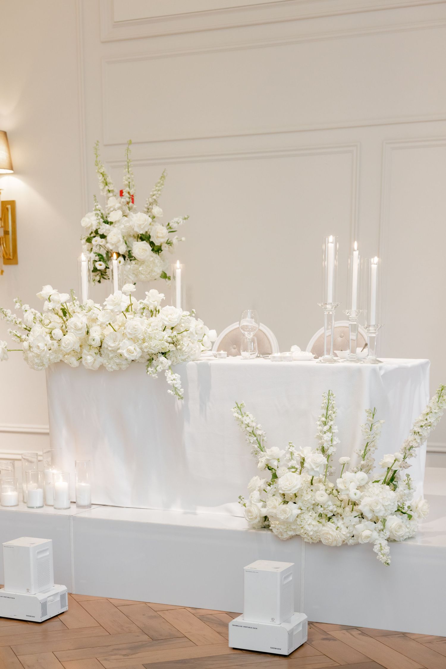 Luxurious white wedding head table with tall candles and fresh floral arrangements on elevated platform.