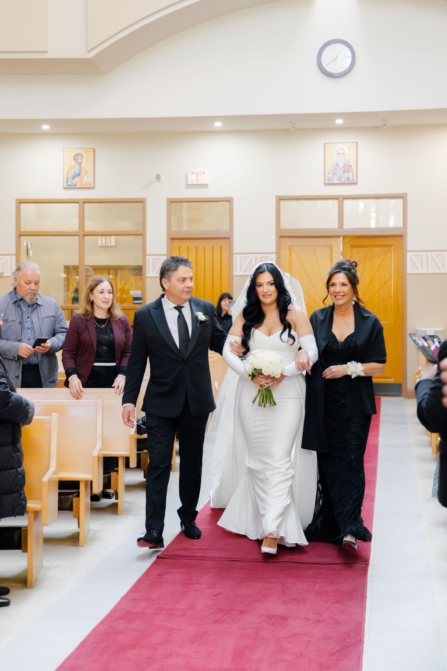 A bridal procession walks down a church aisle with religious artwork on the walls.