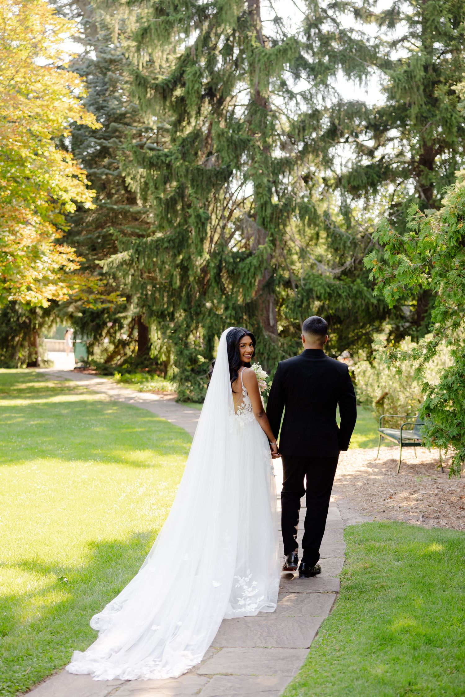 A couple strolls hand in hand down a garden path with trailing lace wedding dress.