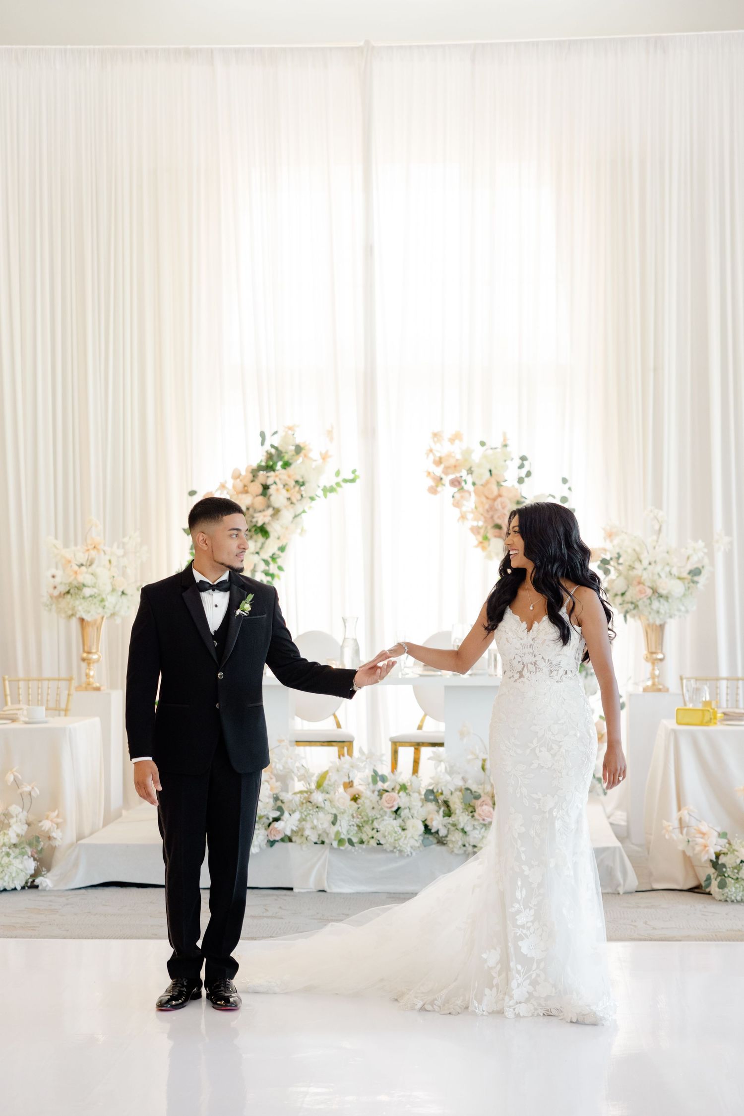 Wedding couple holding hands in front of white backdrop with floral arrangements.