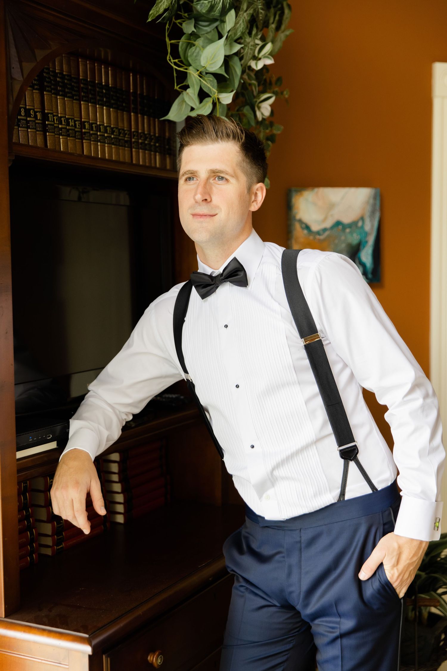 Formal portrait of groomsman in white shirt, suspenders and bow tie against orange wall.