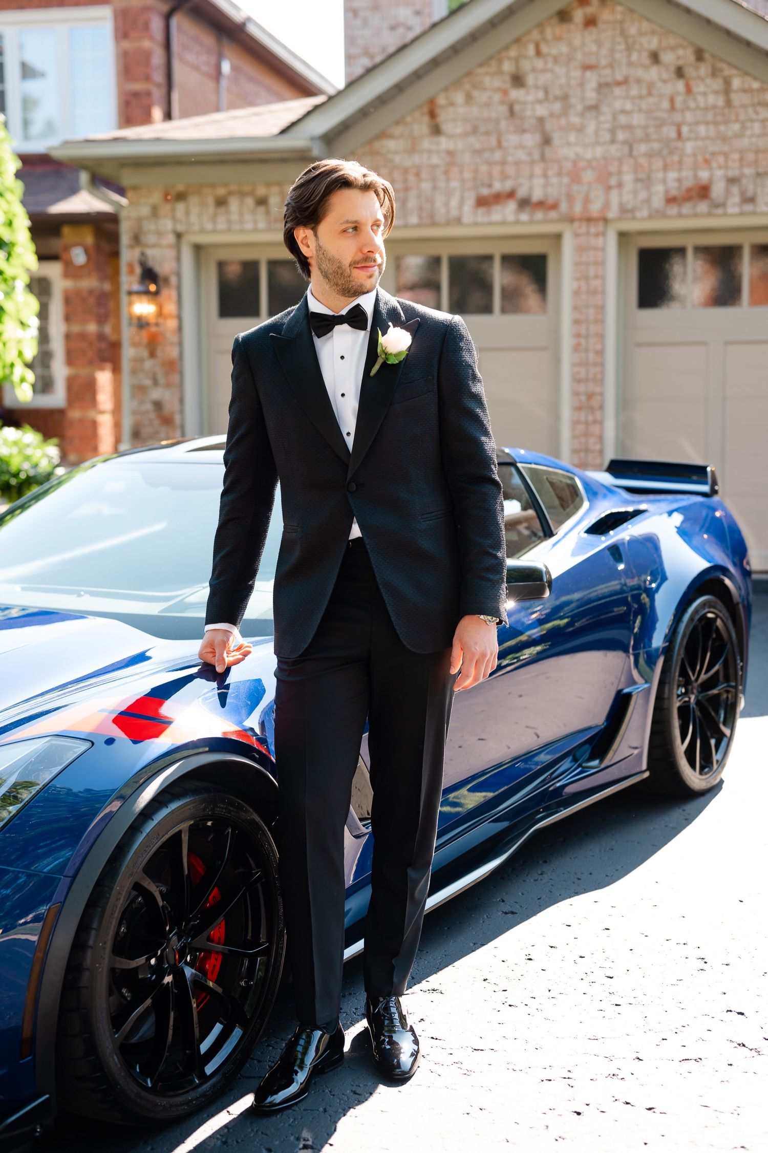 Groom in classic black tuxedo poses next to sleek blue sports car outside brick house.