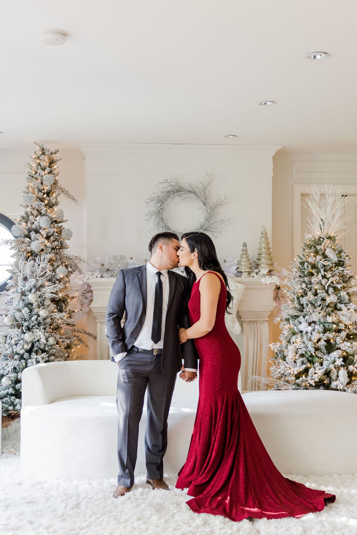 Romantic formal portrait in front of decorated Christmas trees with red gown and gray suit.