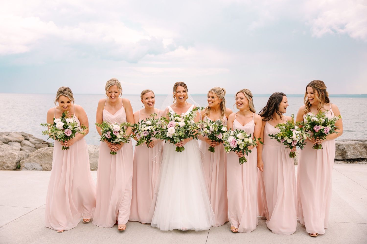 Wedding party holding white floral bouquets pose together on a stone waterfront.