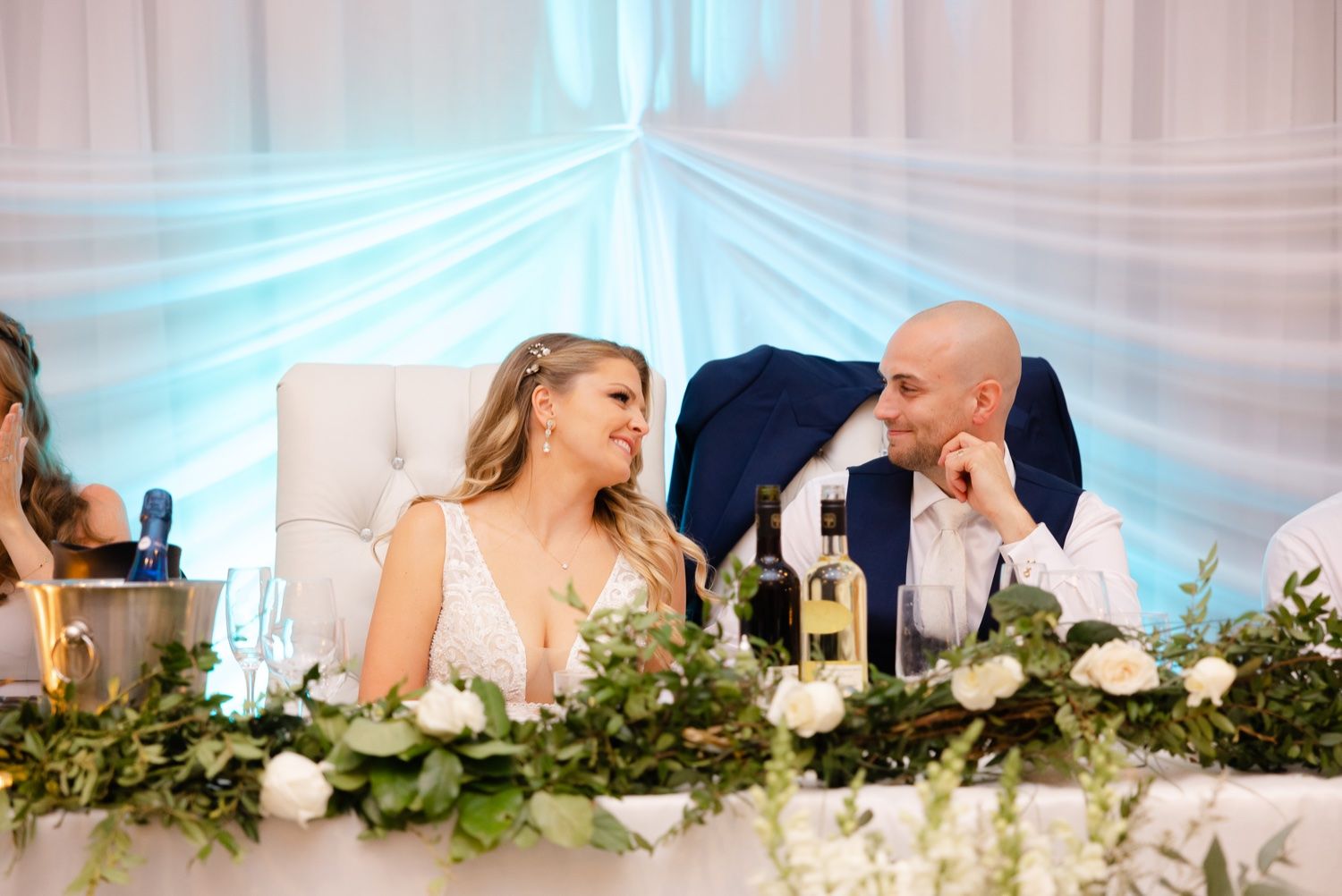 Couple sitting at head table decorated with greenery garland at wedding reception.