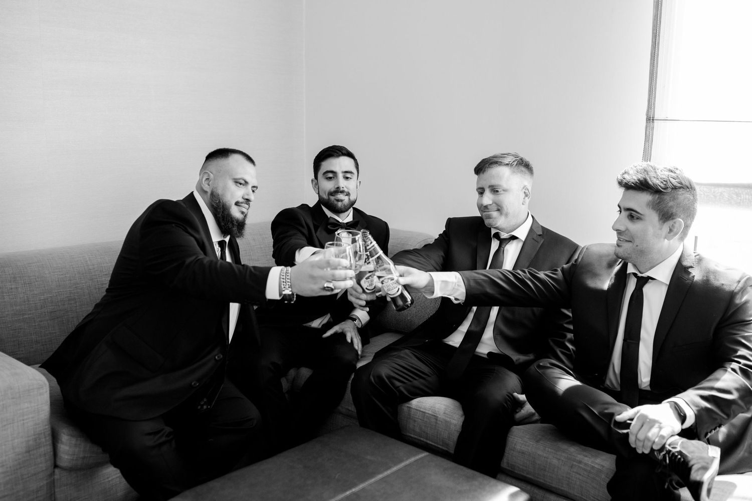 Groomsmen in black suits sharing drinks while sitting on a couch during wedding preparations.