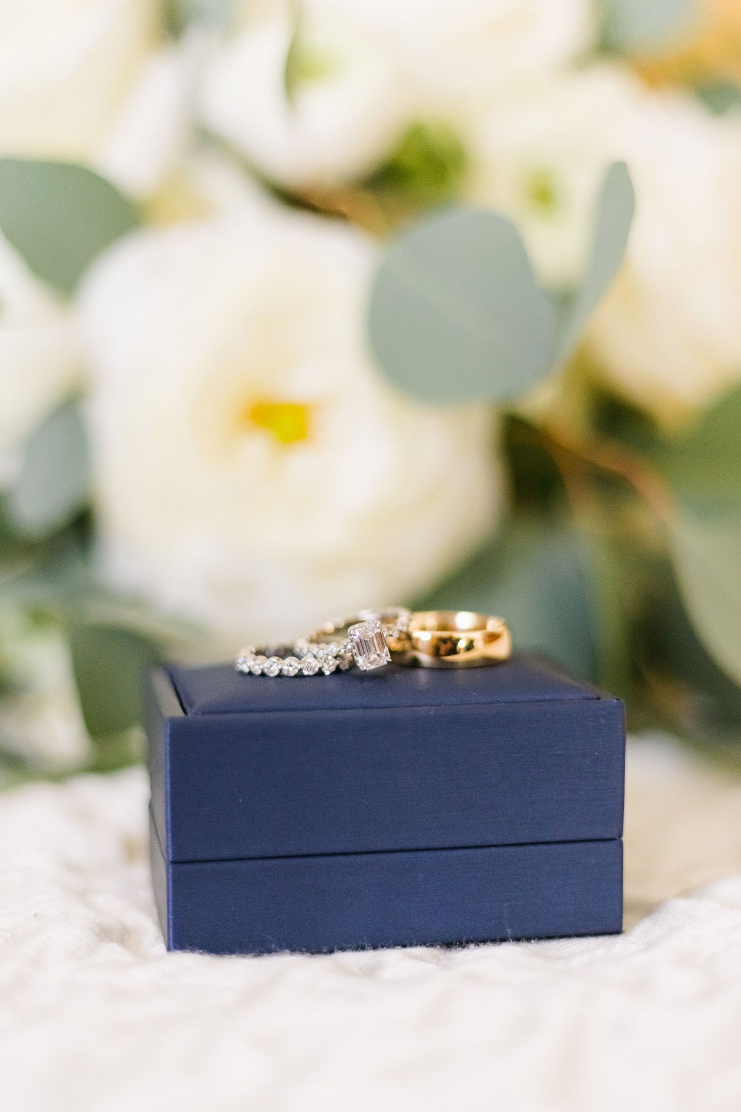Wedding rings displayed on navy blue box with white flowers and eucalyptus in background.