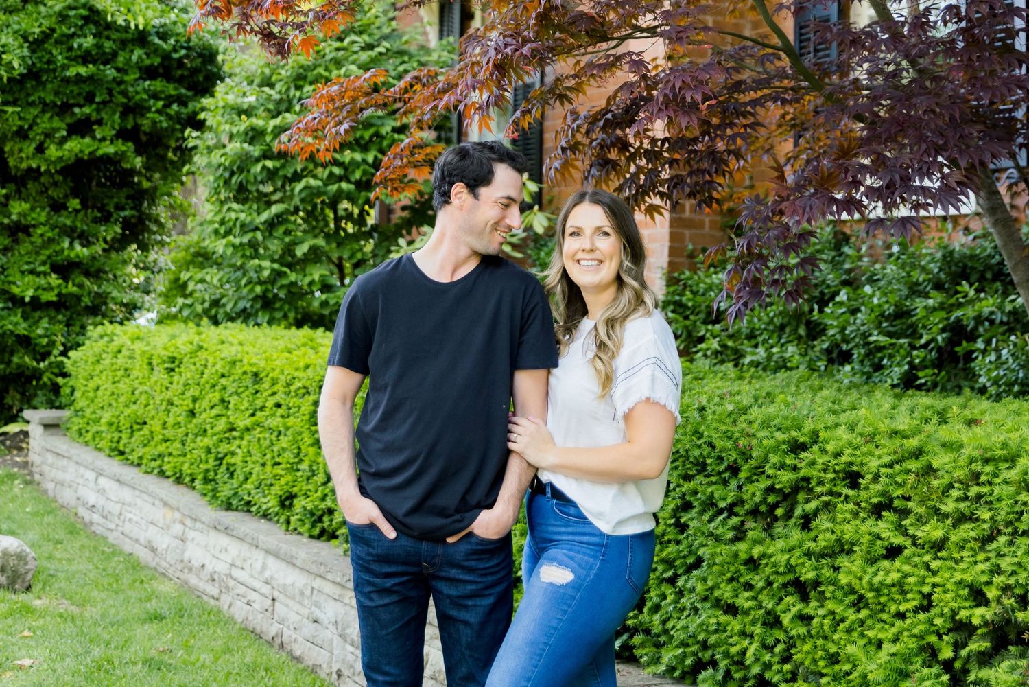 A couple smiles together against lush green landscaping and a dramatic red maple tree.