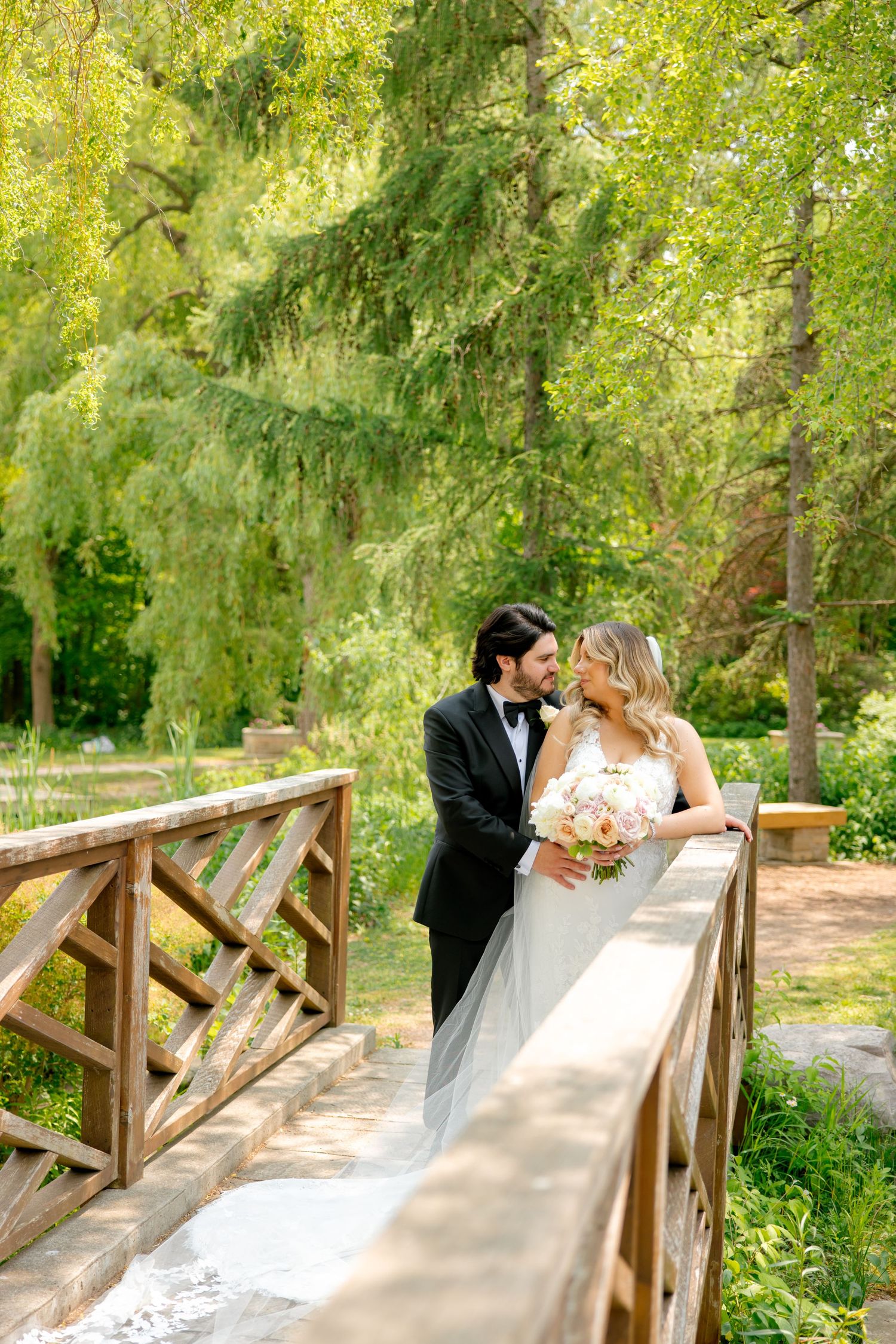 Elegant wedding couple posing on rustic wooden bridge surrounded by lush greenery.