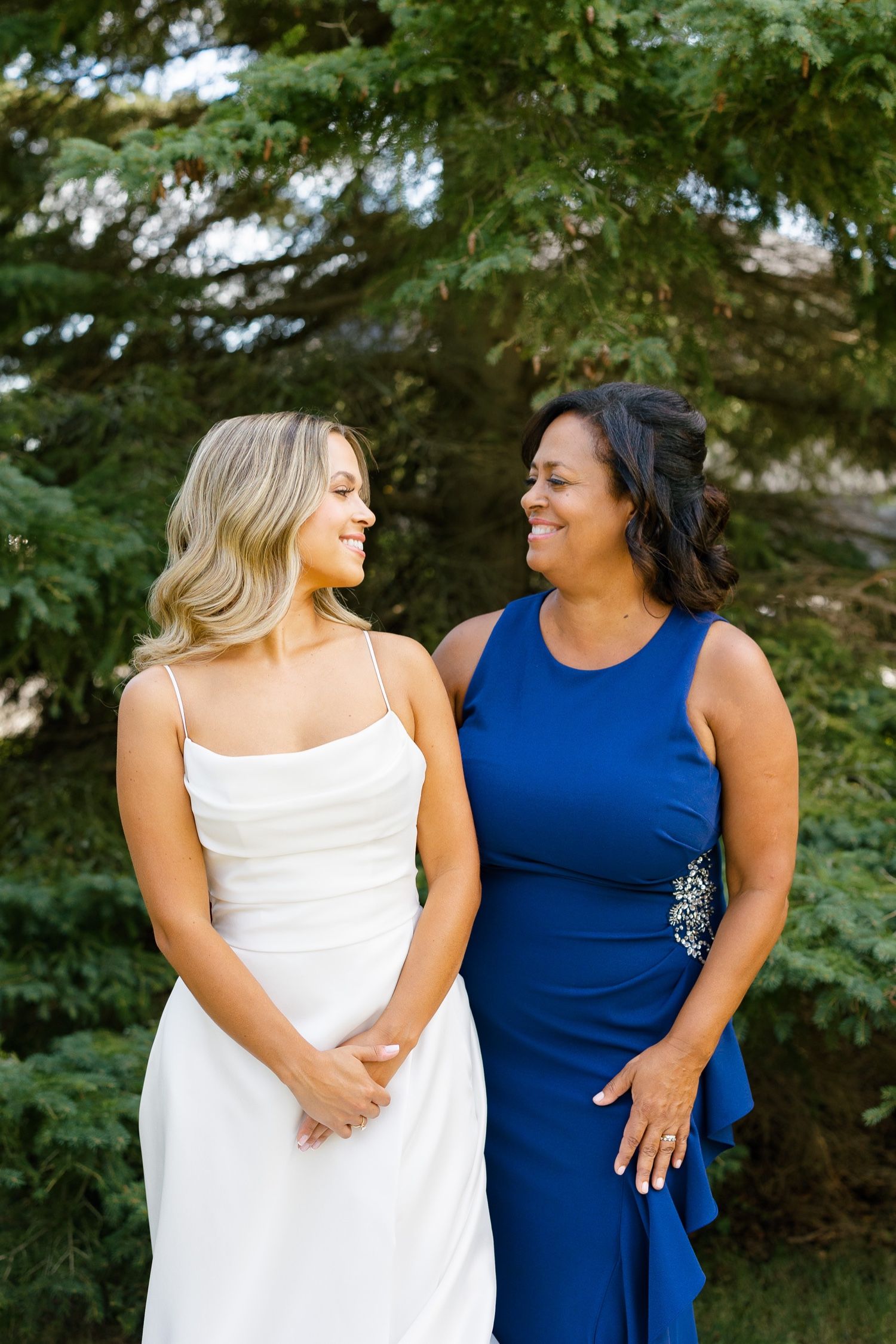 Two people in formal attire share a joyful moment outdoors in front of evergreen trees.