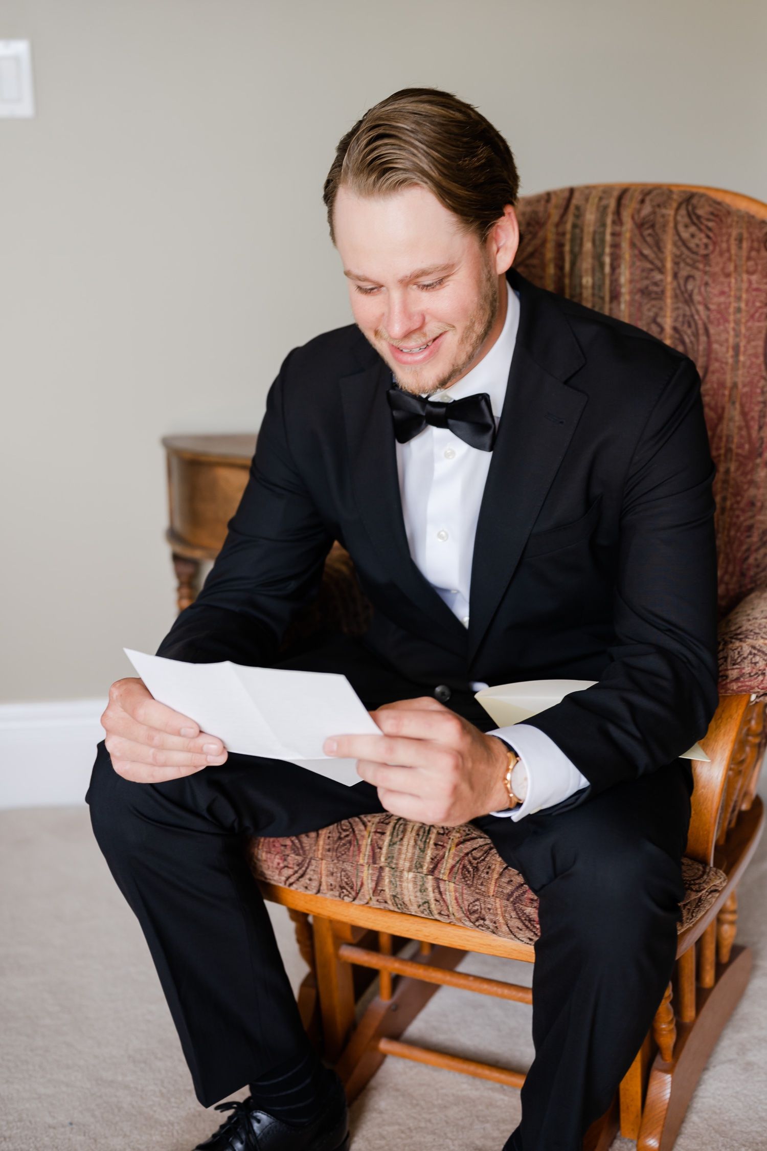 Person in black tuxedo sitting in wooden rocking chair reading documents with a gentle smile.