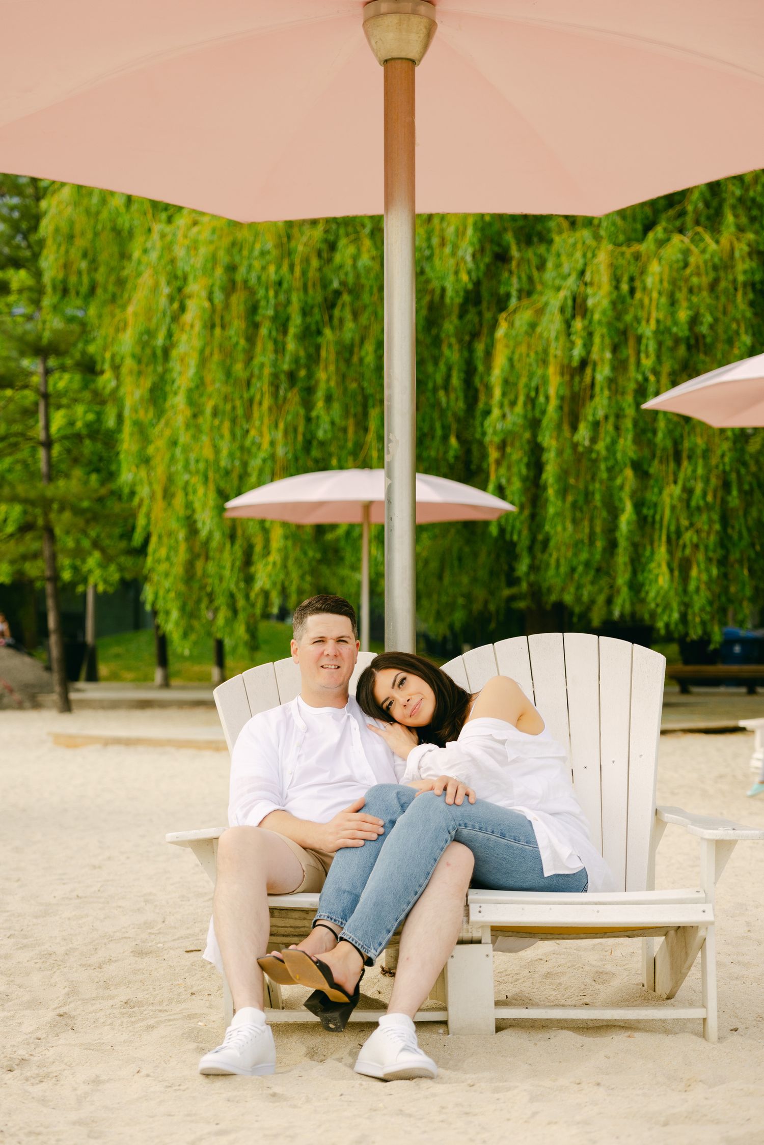 A couple relaxes together on an outdoor lounge chair under a pink umbrella.