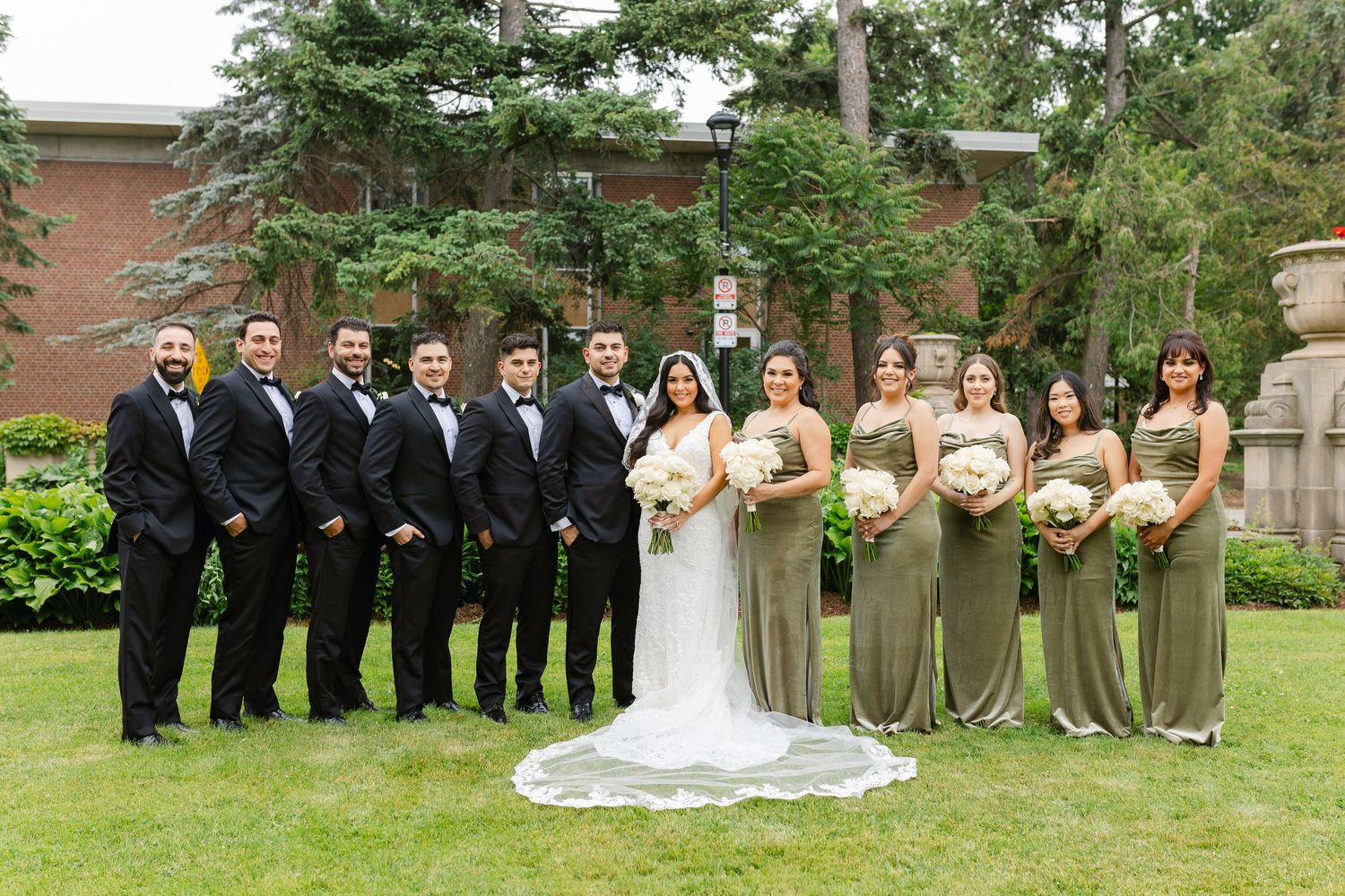 Wedding party poses outdoors with bridesmaids in sage dresses and groomsmen in black tuxedos.
