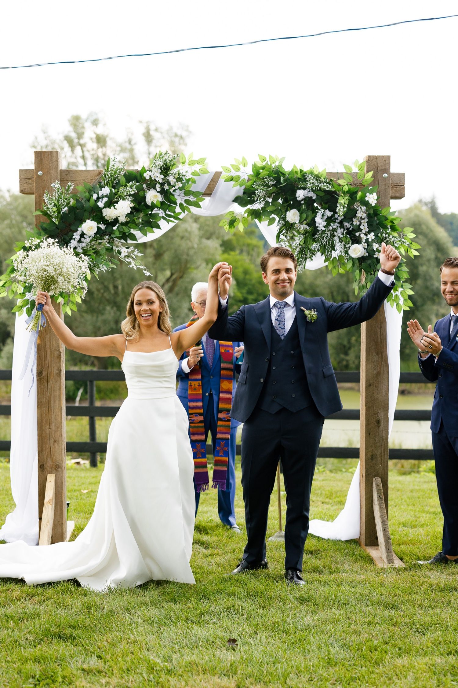 Newlyweds celebrate with raised arms under a wooden arch adorned with white flowers after their ceremony.