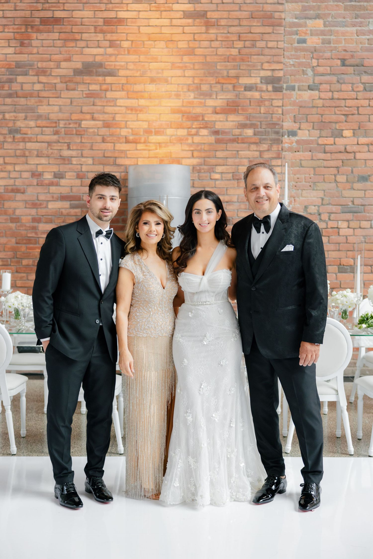 Four people in formal attire pose together at elegant wedding venue with brick wall backdrop.