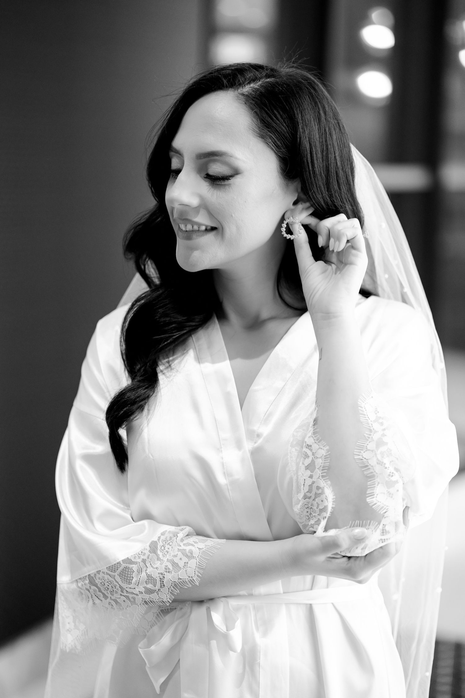 Bride in white lace robe adjusts earring while getting ready for wedding day.