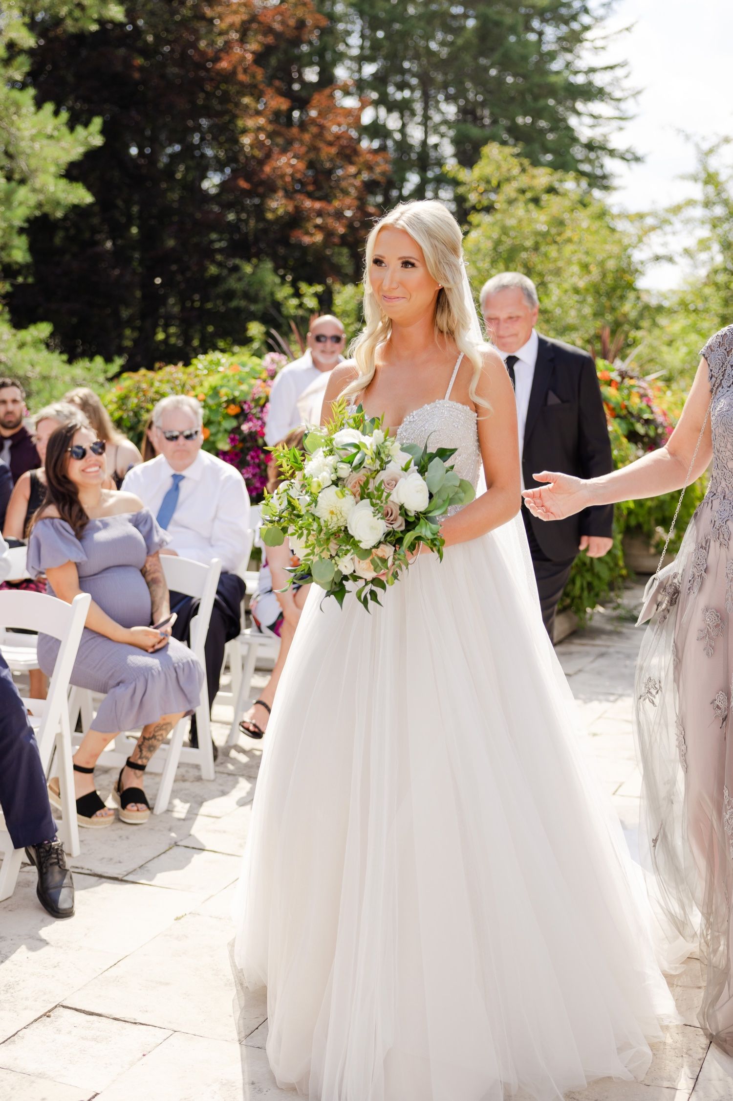 A bride in a white ballgown carries a fresh bouquet during an outdoor garden wedding ceremony.