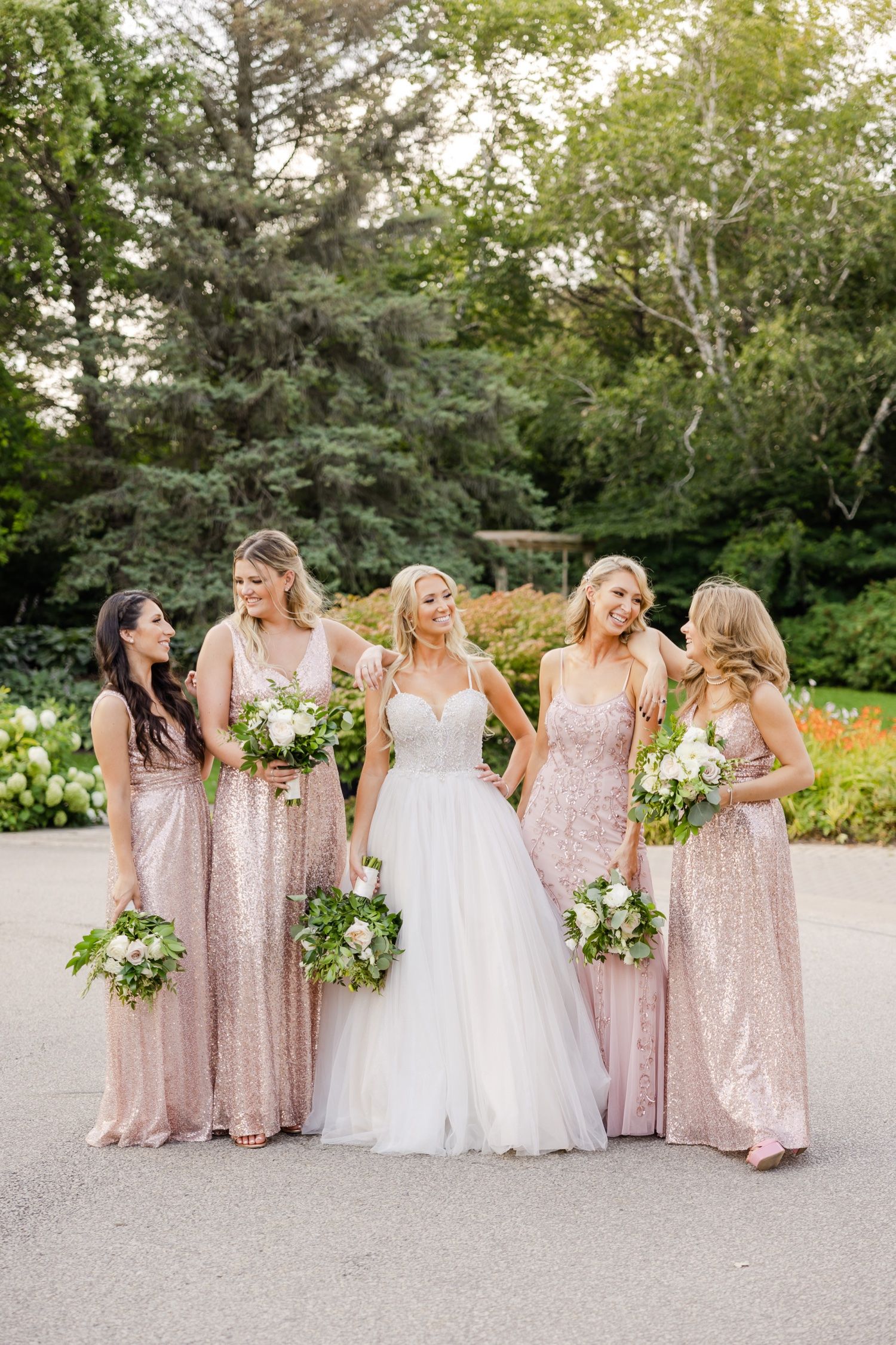Bridal party in sparkly pink dresses poses with bride in white gown on garden path.