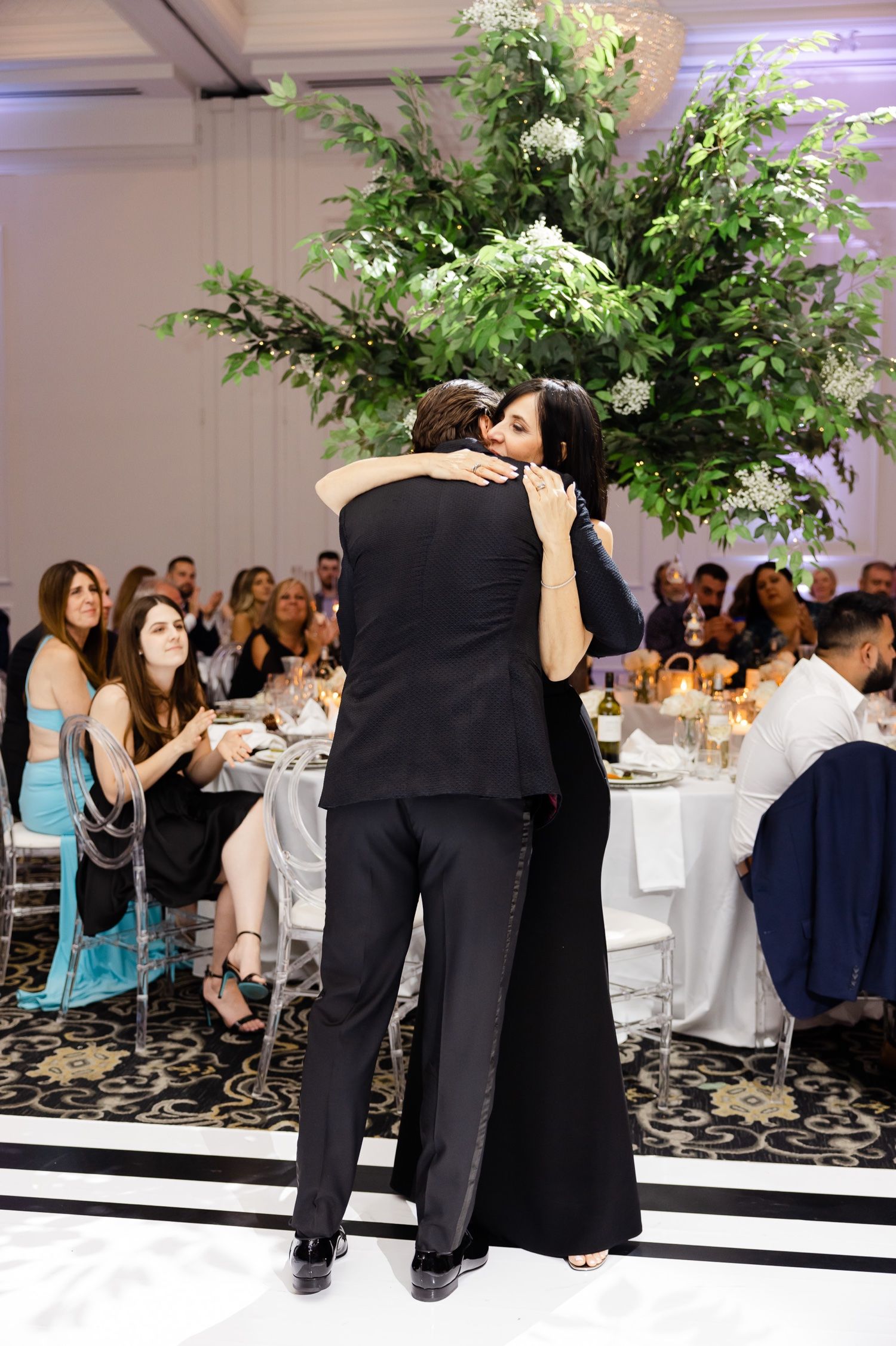 Couple embracing on dance floor as wedding guests look on from decorated reception tables.
