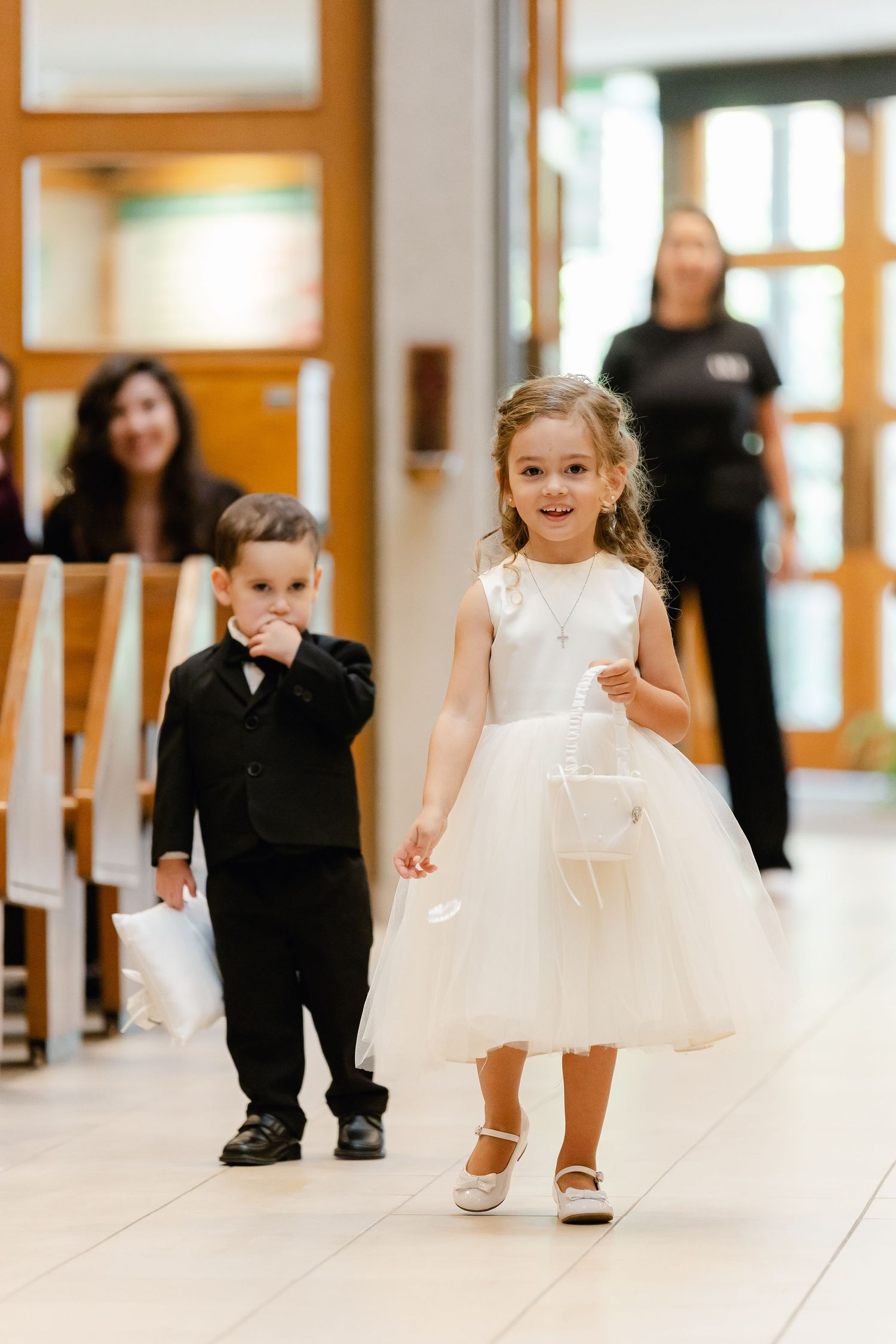 Ring bearer in black suit and flower girl in white dress walk down church aisle together.
