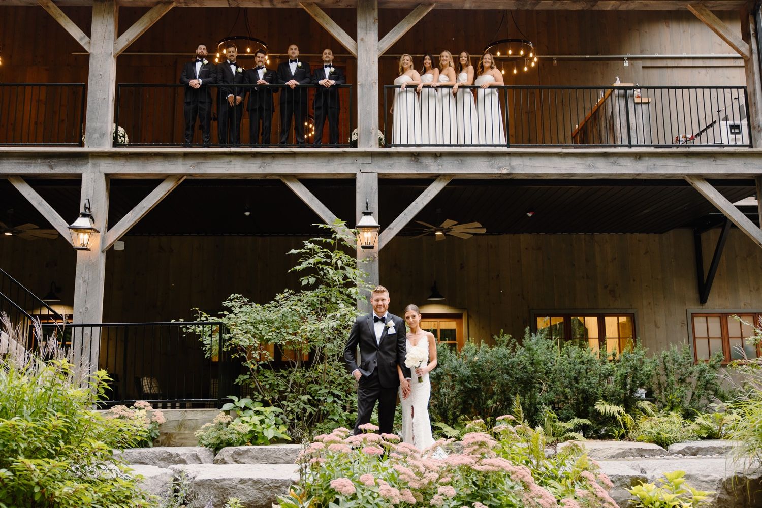 Wedding couple poses in front of rustic venue with bridal party watching from two-story balcony.