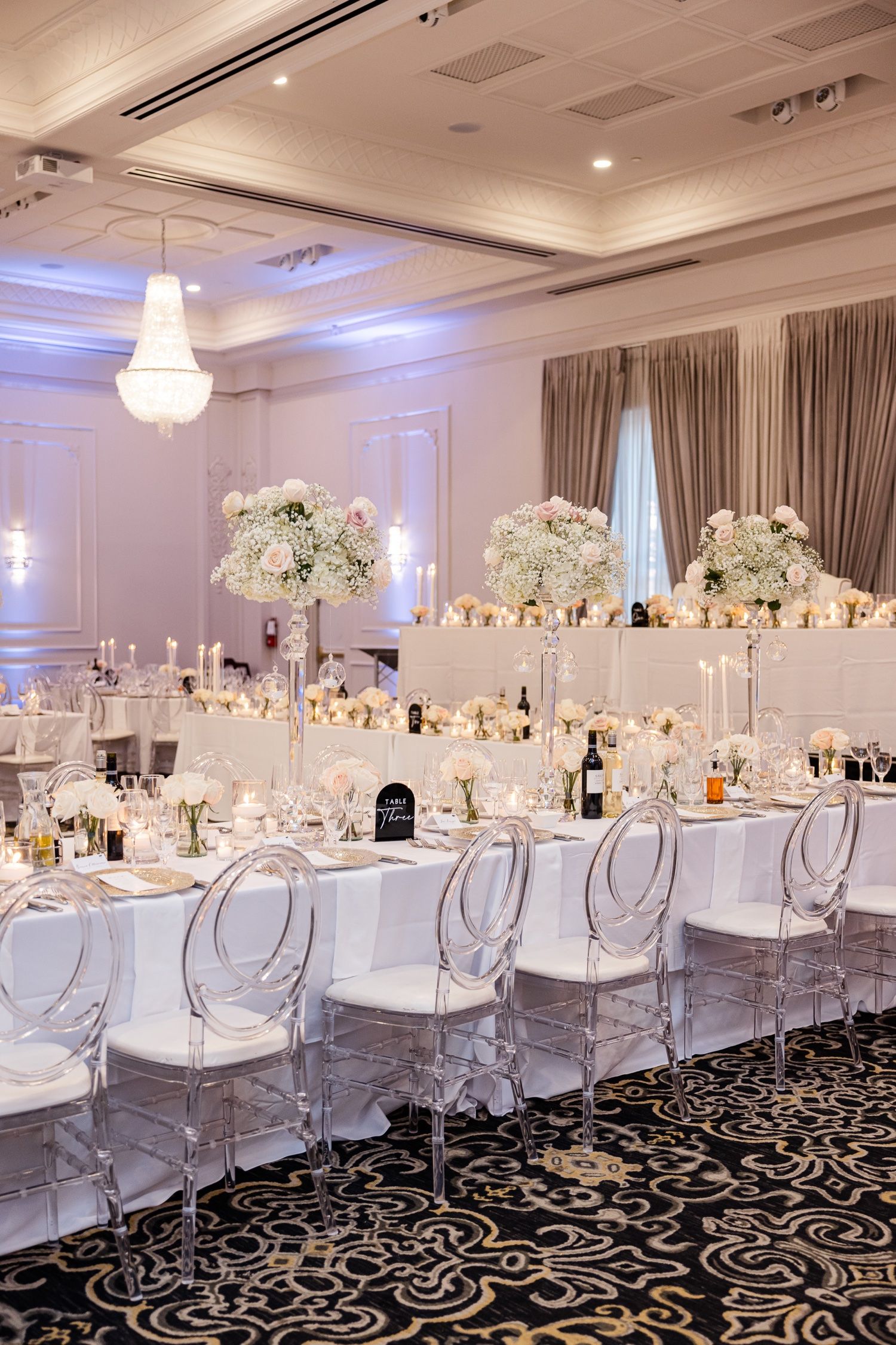 Luxurious wedding reception with crystal chandelier, ghost chairs, and white floral centerpieces.