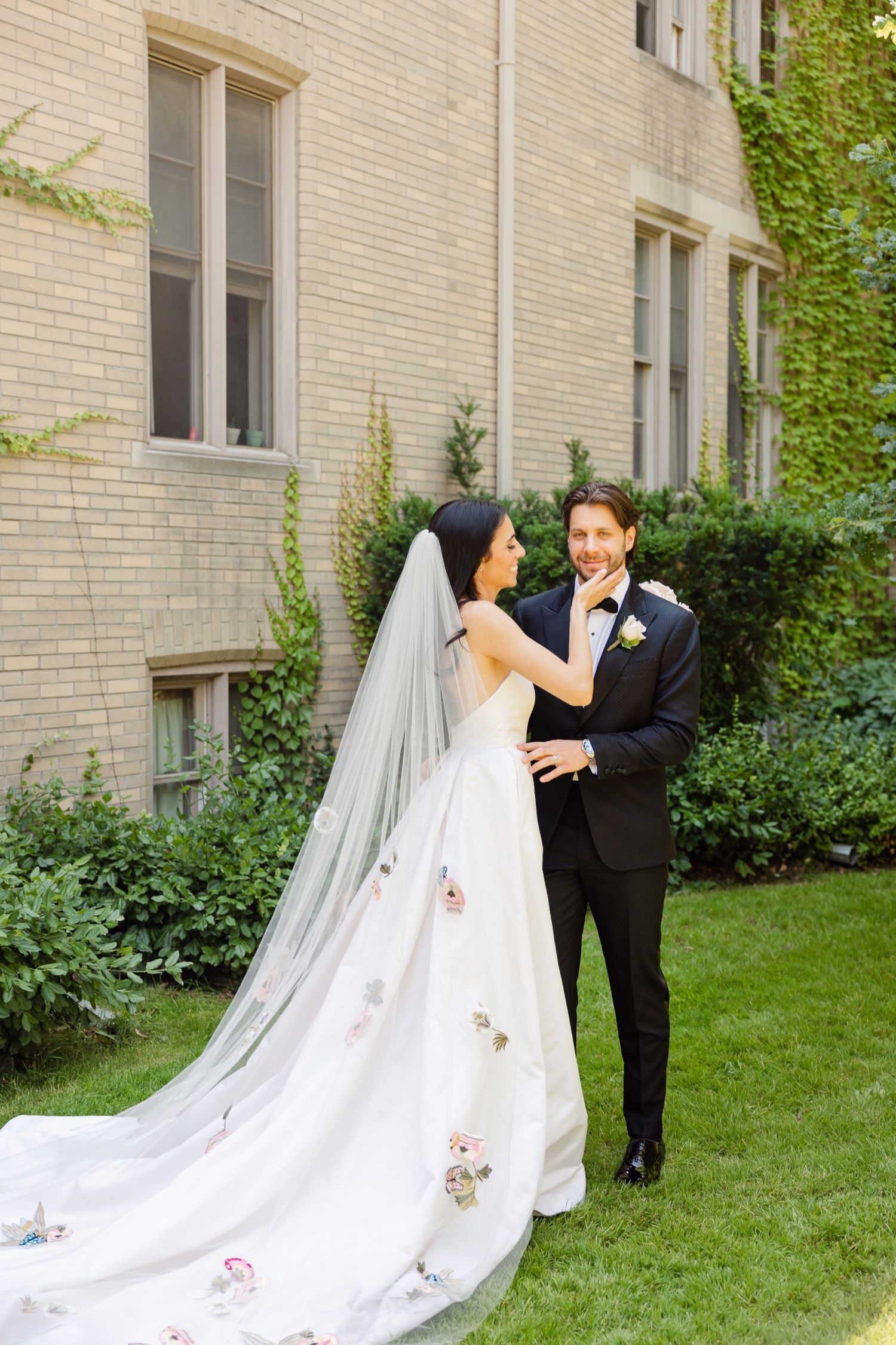A romantic wedding photo showing a couple embracing on a manicured lawn with an ivy-covered backdrop.