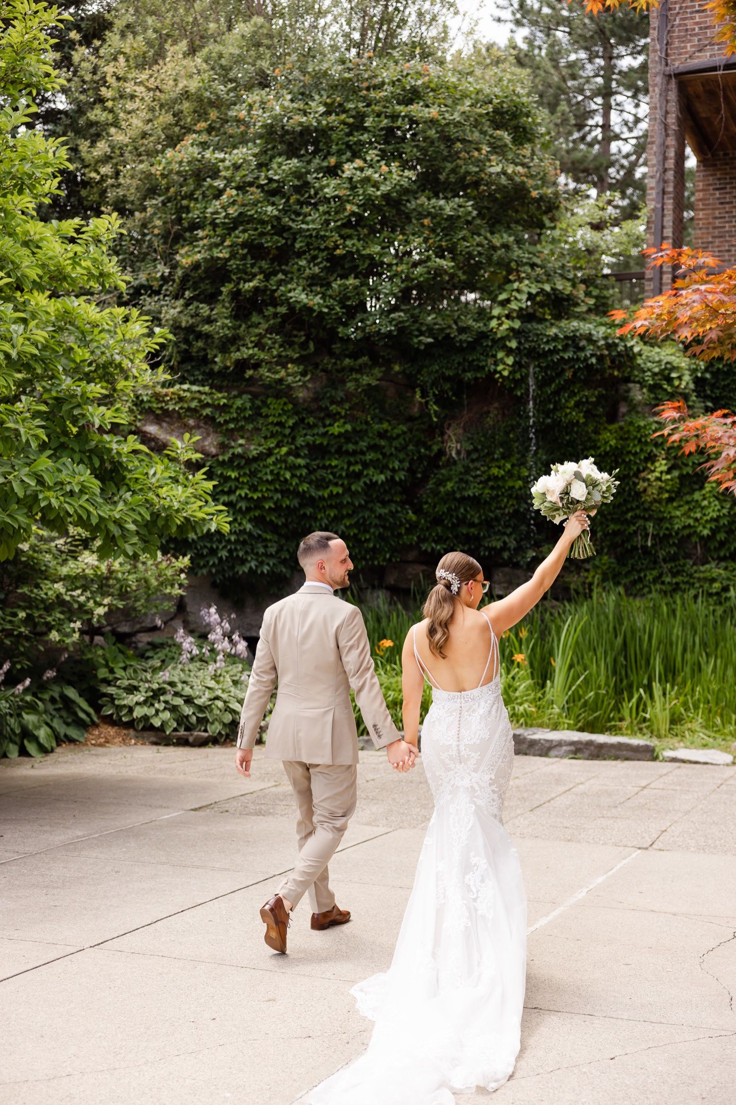 Newlyweds walk hand in hand through a lush garden path with bride holding white bouquet.