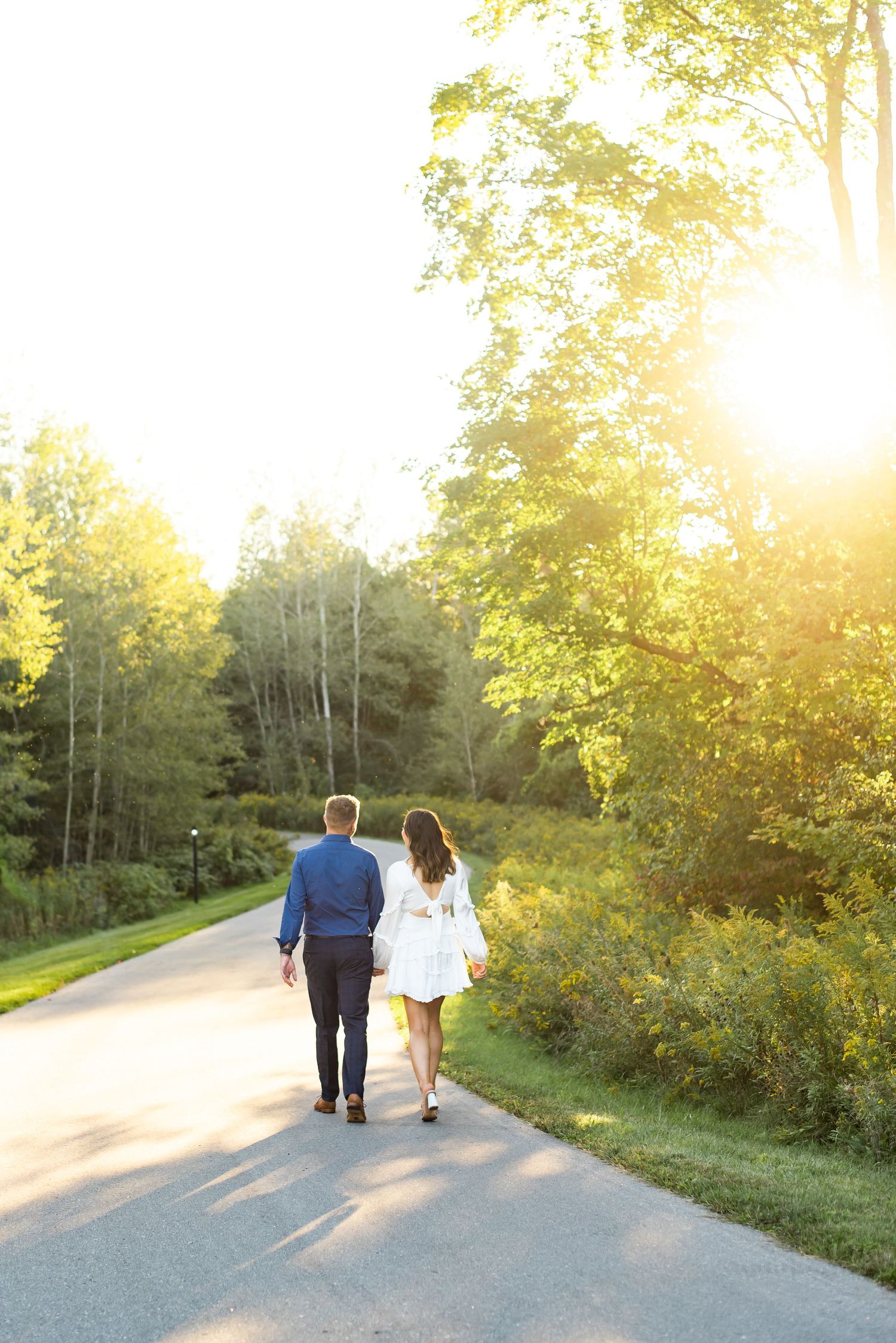 A couple walks hand in hand down a sunlit path surrounded by lush summer greenery.