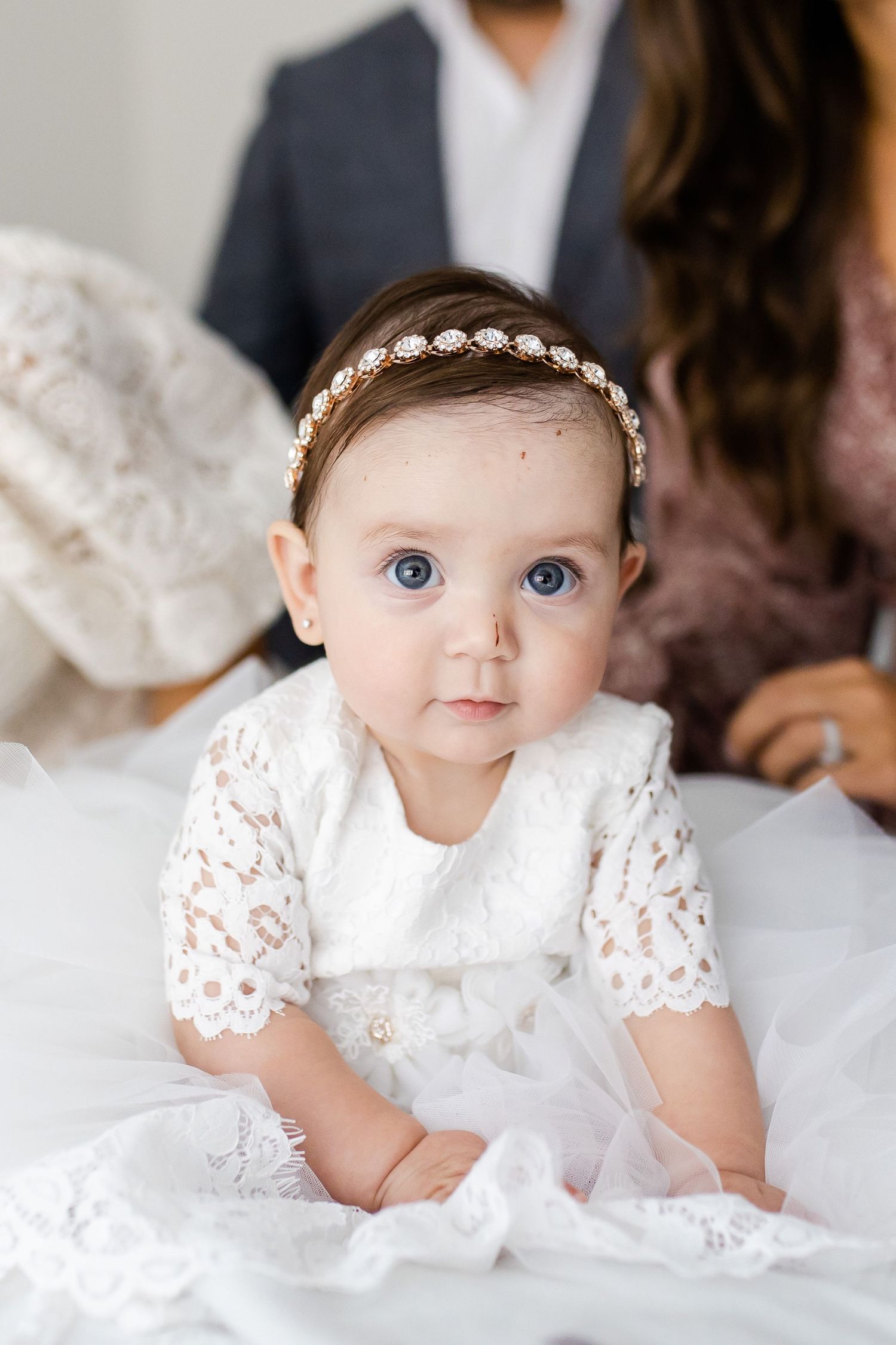 Close up of baby in white lace dress wearing delicate pearl headband on white bedding.