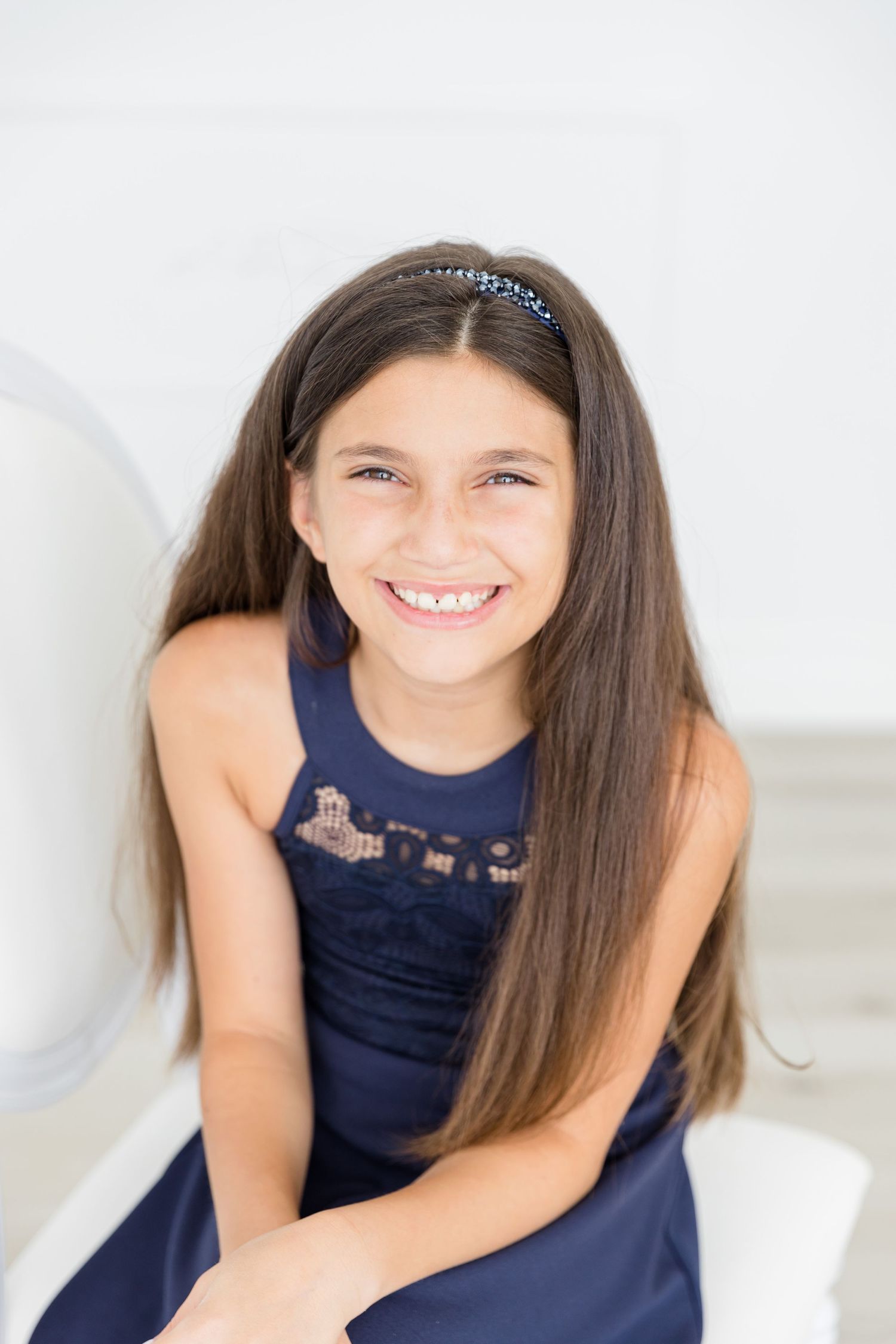 Girl in navy dress smiles brightly while sitting against white background.