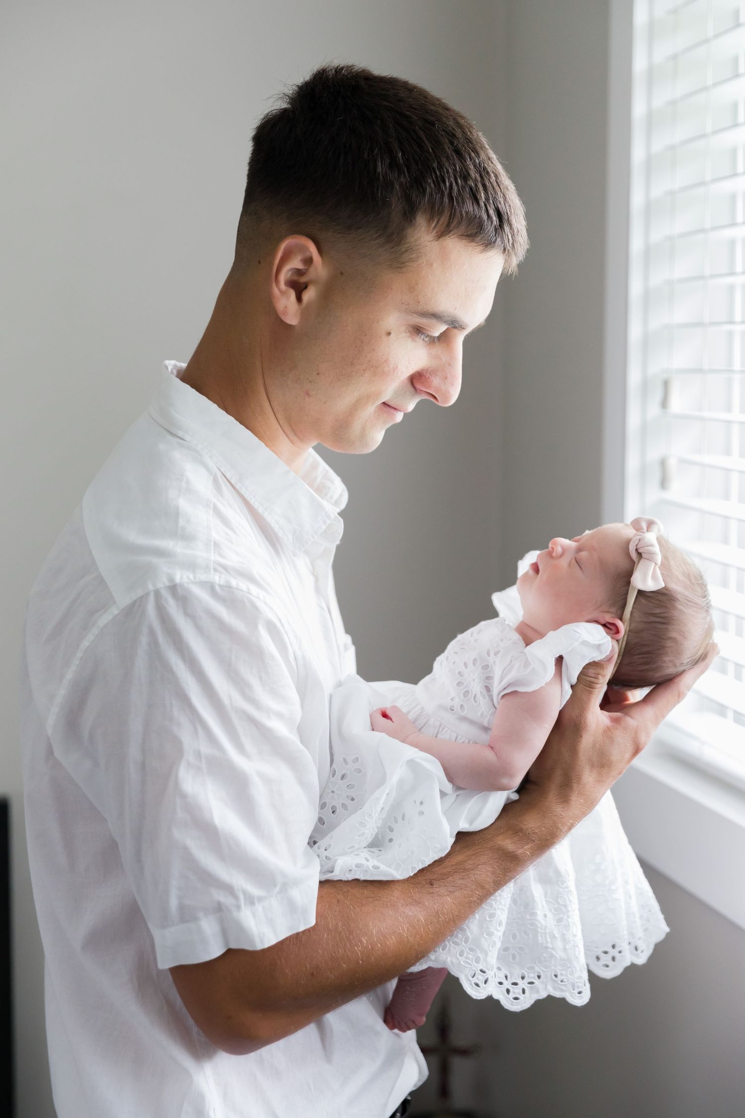Parent in white shirt holding newborn baby dressed in white lace near window with natural lighting.